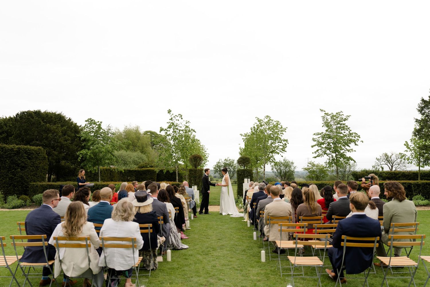 Guests seated on wooden chairs at an outdoor garden wedding ceremony on a lush green lawn.