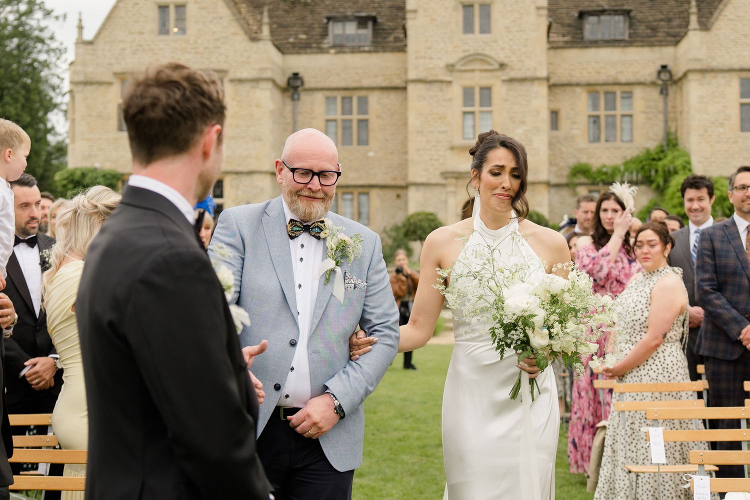 Bride in halter neck gown walks down outdoor aisle with escort at a grand English country house wedding.