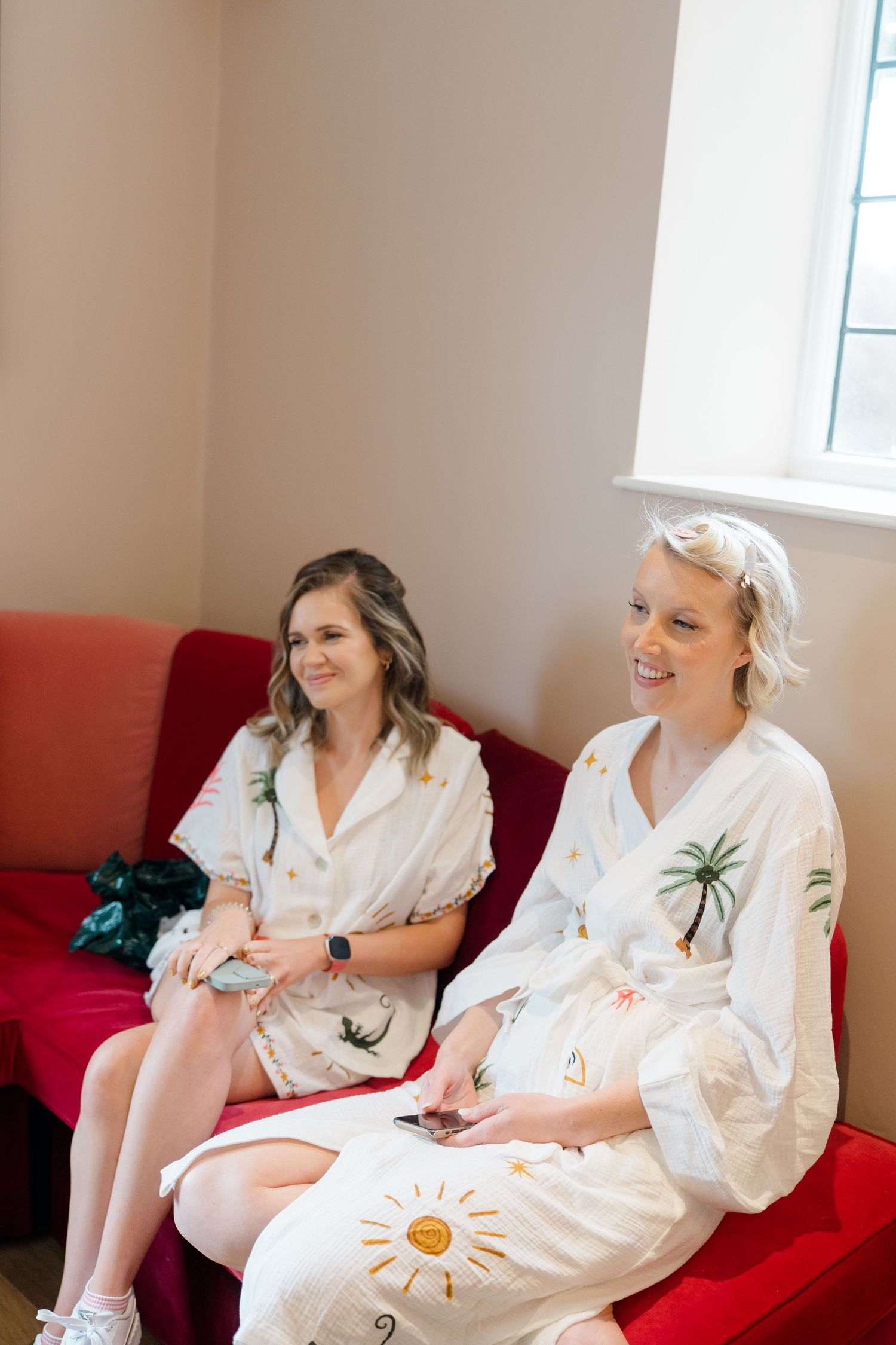 Two women in matching white robes sit on a red sofa, smiling and relaxing before a wedding ceremony.