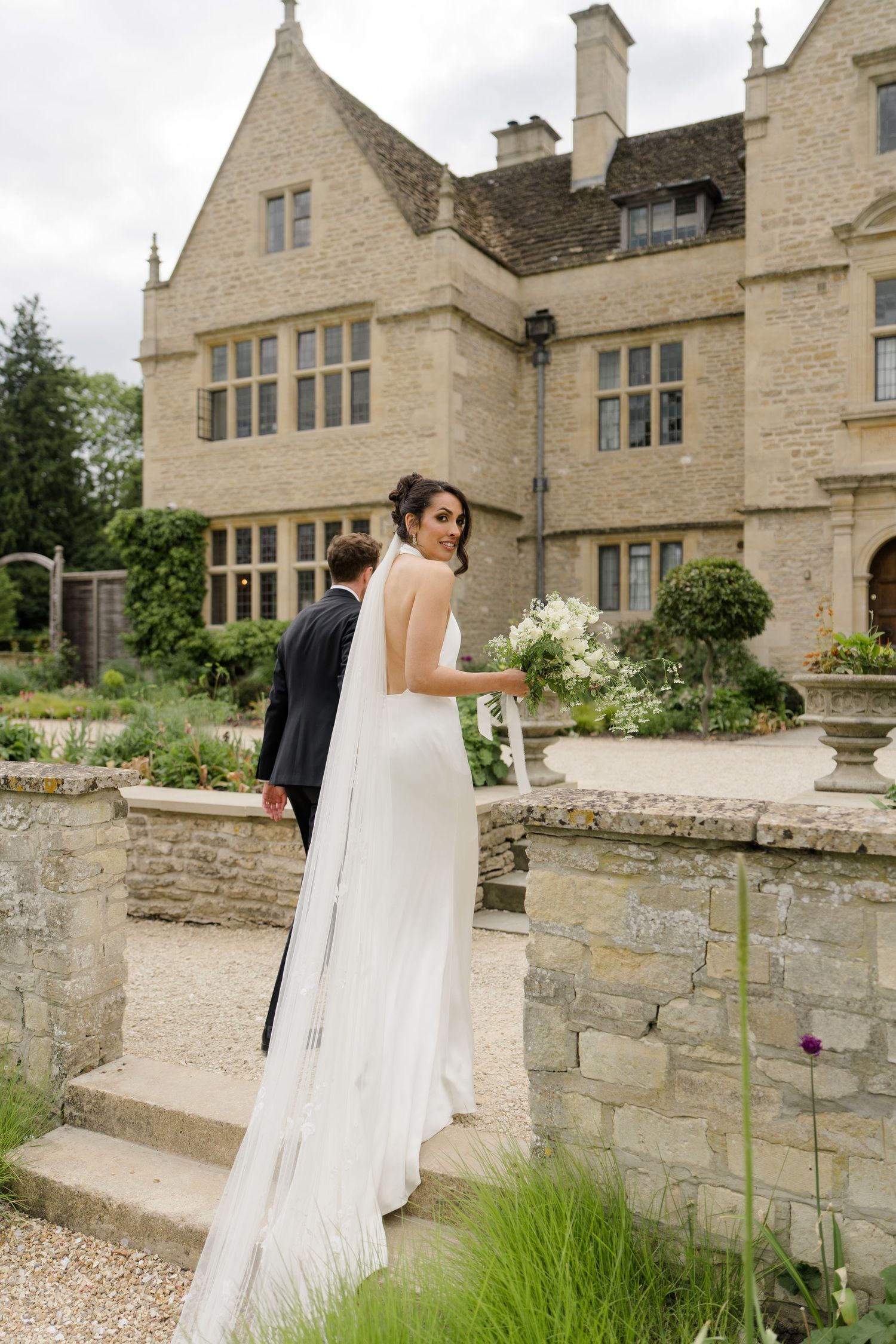 Bride in backless white gown with cathedral veil poses with groom on stone steps of an English manor.