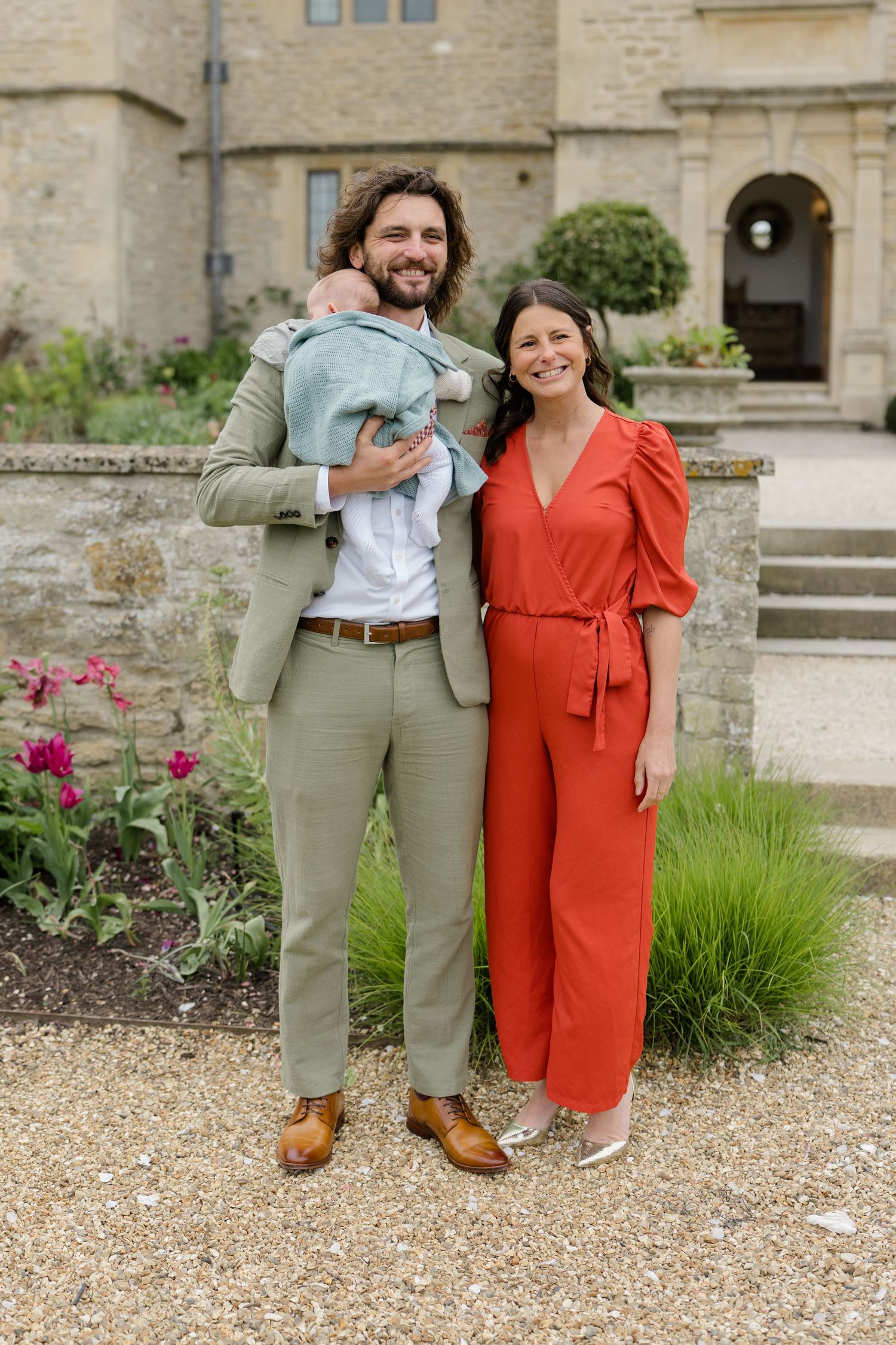 Smiling man in sage suit holding a baby beside a woman in a red jumpsuit outside a stone manor.