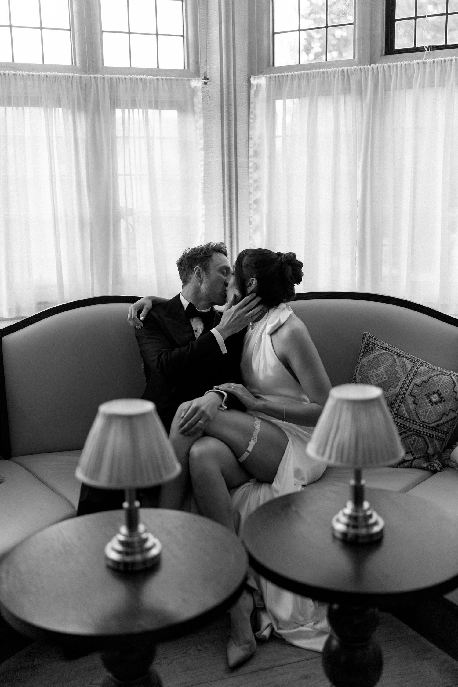 Black and white photo of a bride and groom sharing a kiss on a sofa by large curtained windows.