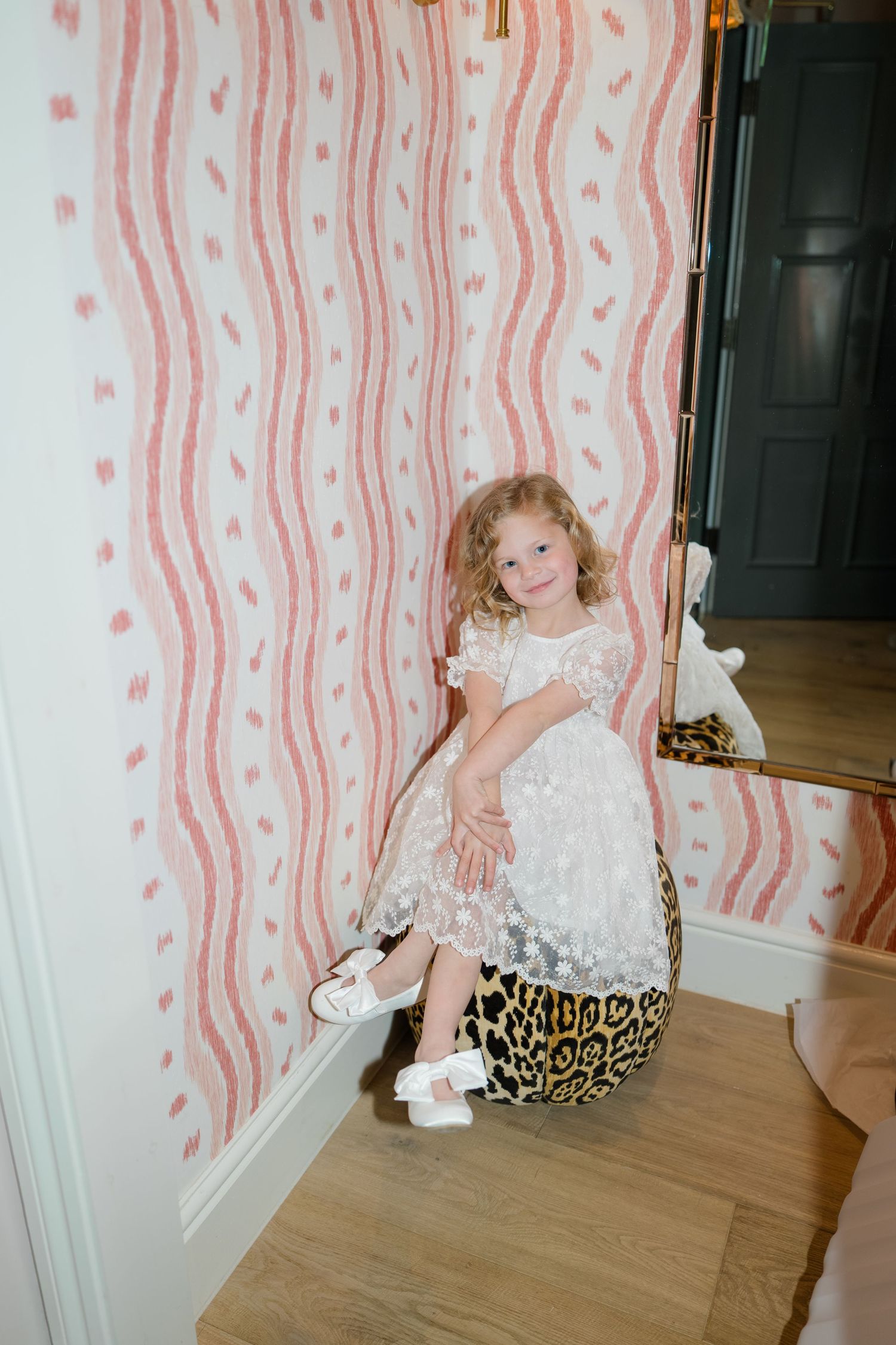 Young flower girl in white lace dress sits on leopard print stool against pink striped wallpaper.