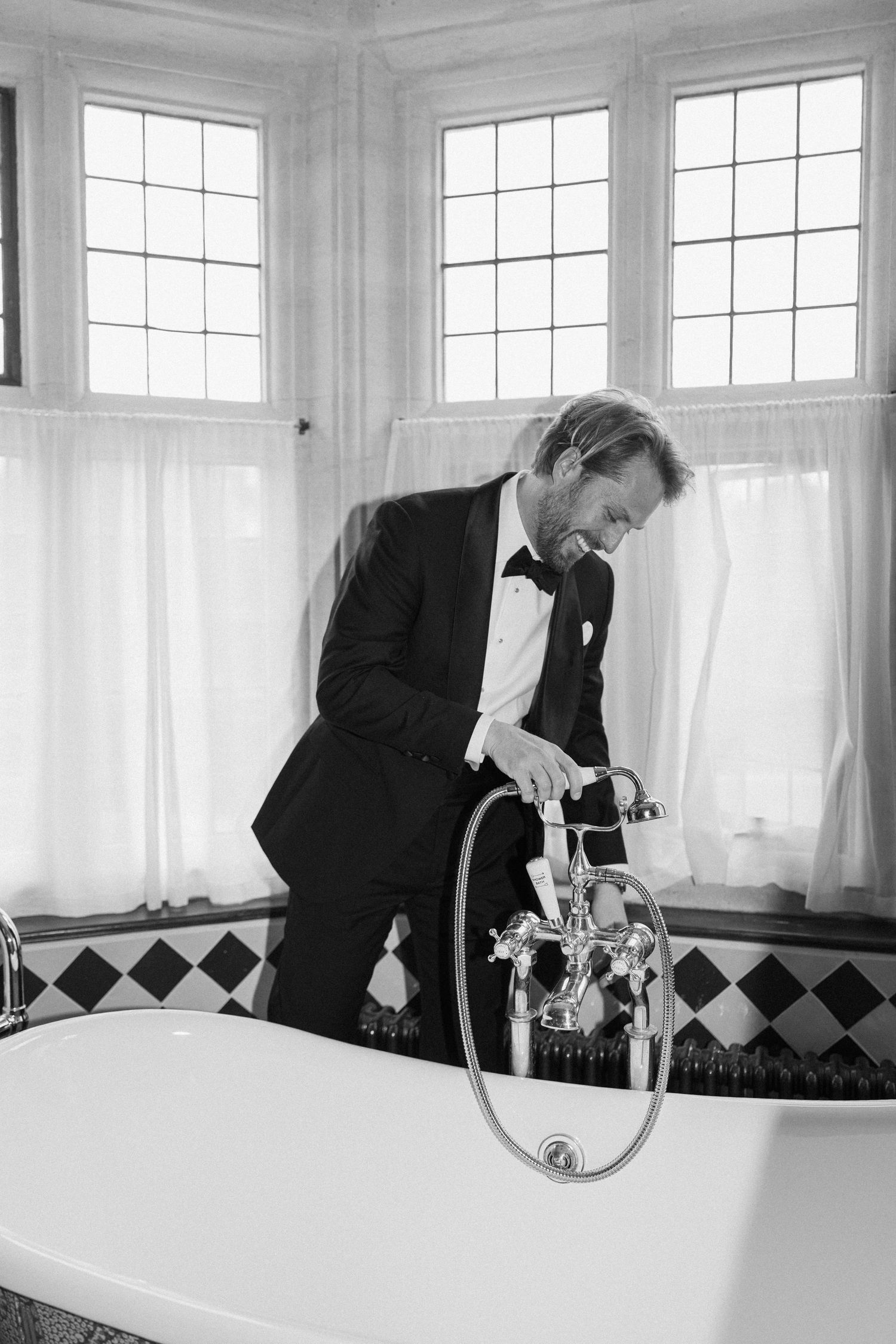 Groom in tuxedo leaning over elegant freestanding bathtub in black and white tiled bathroom.