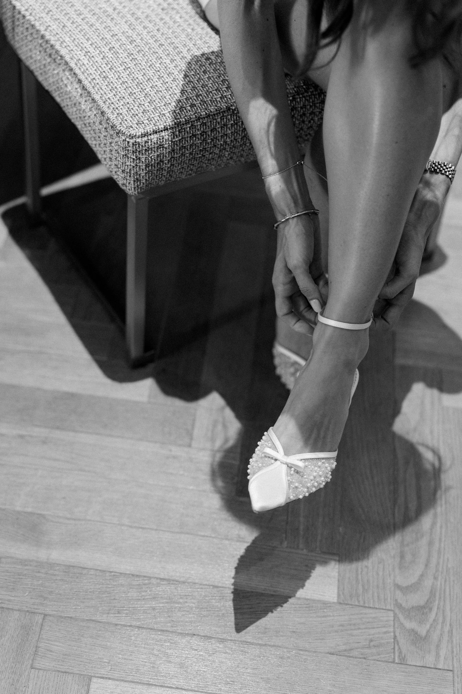 Black and white photo of a bride putting on white heeled wedding shoes while seated on a bench.