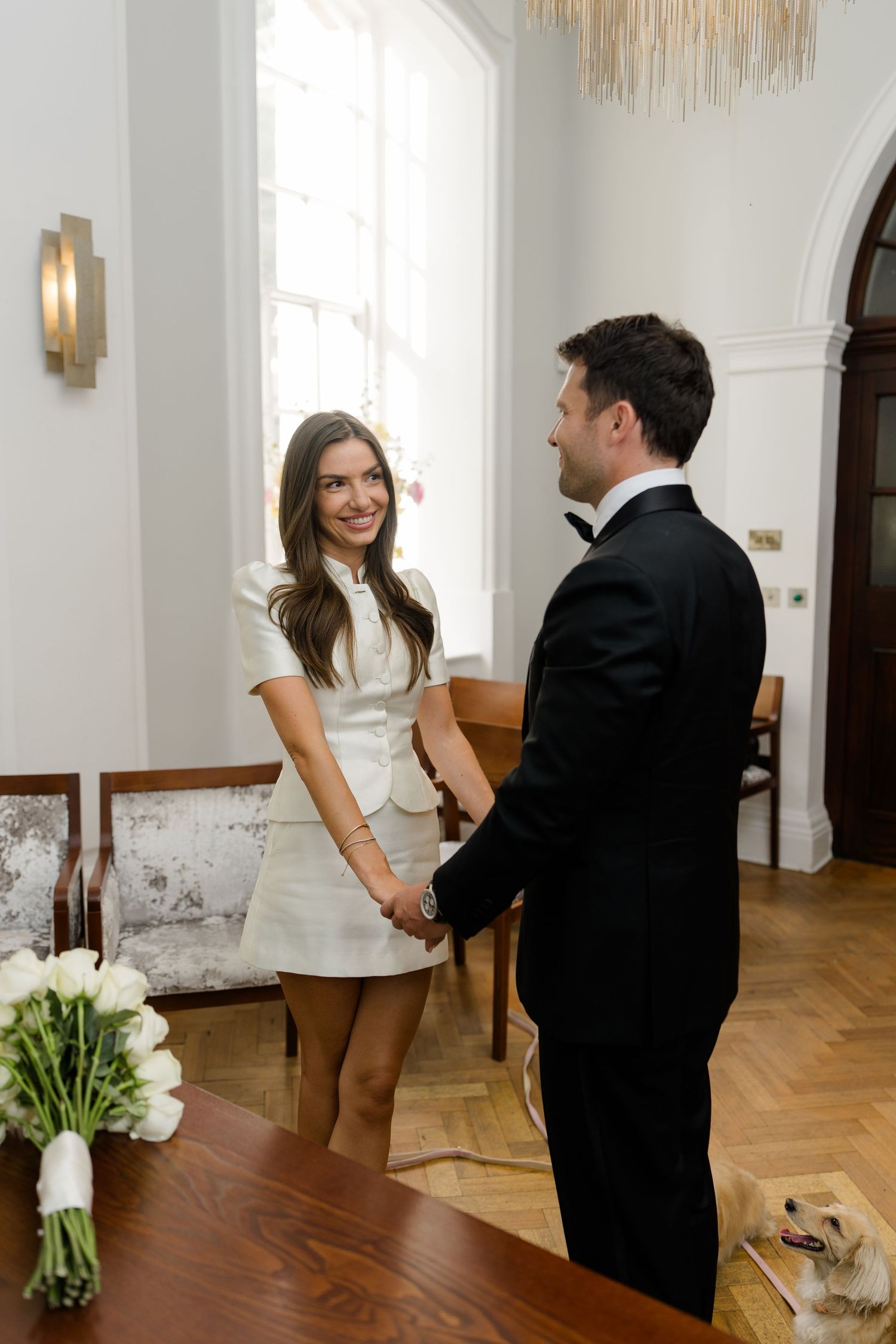 Bride in white mini dress and groom in tuxedo holding hands at a bright registry office.
