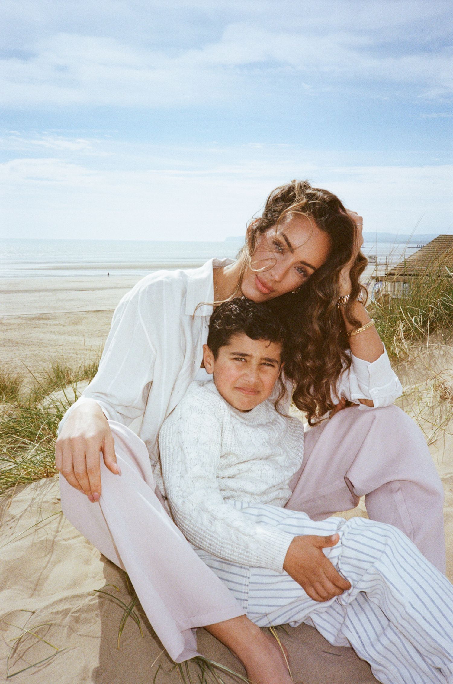 Woman and young boy sitting together on beach sand dunes, smiling warmly in casual white and pink outfits.