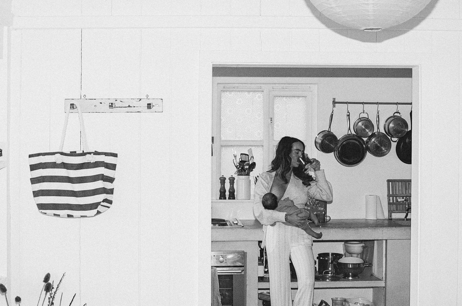 Black and white photo of a woman holding a baby in a cozy kitchen with hanging pots and a striped tote bag.