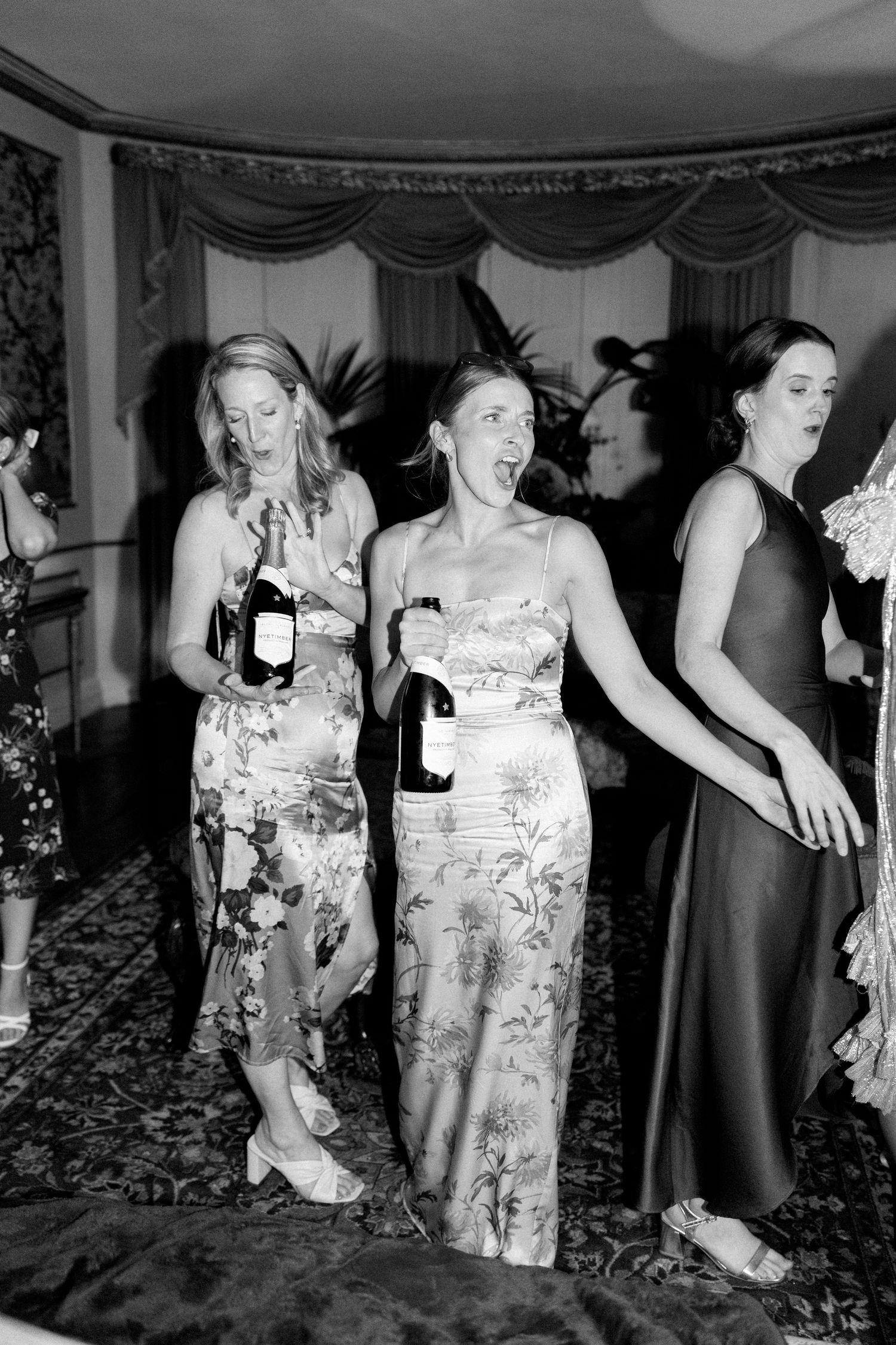 Wedding celebration with attendees in formal attire holding wine bottles and dancing.
