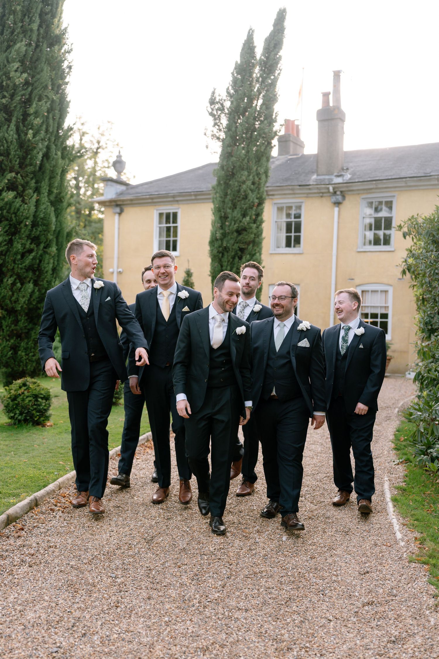 Wedding party in matching dark suits walking together on a gravel path outside a yellow building.