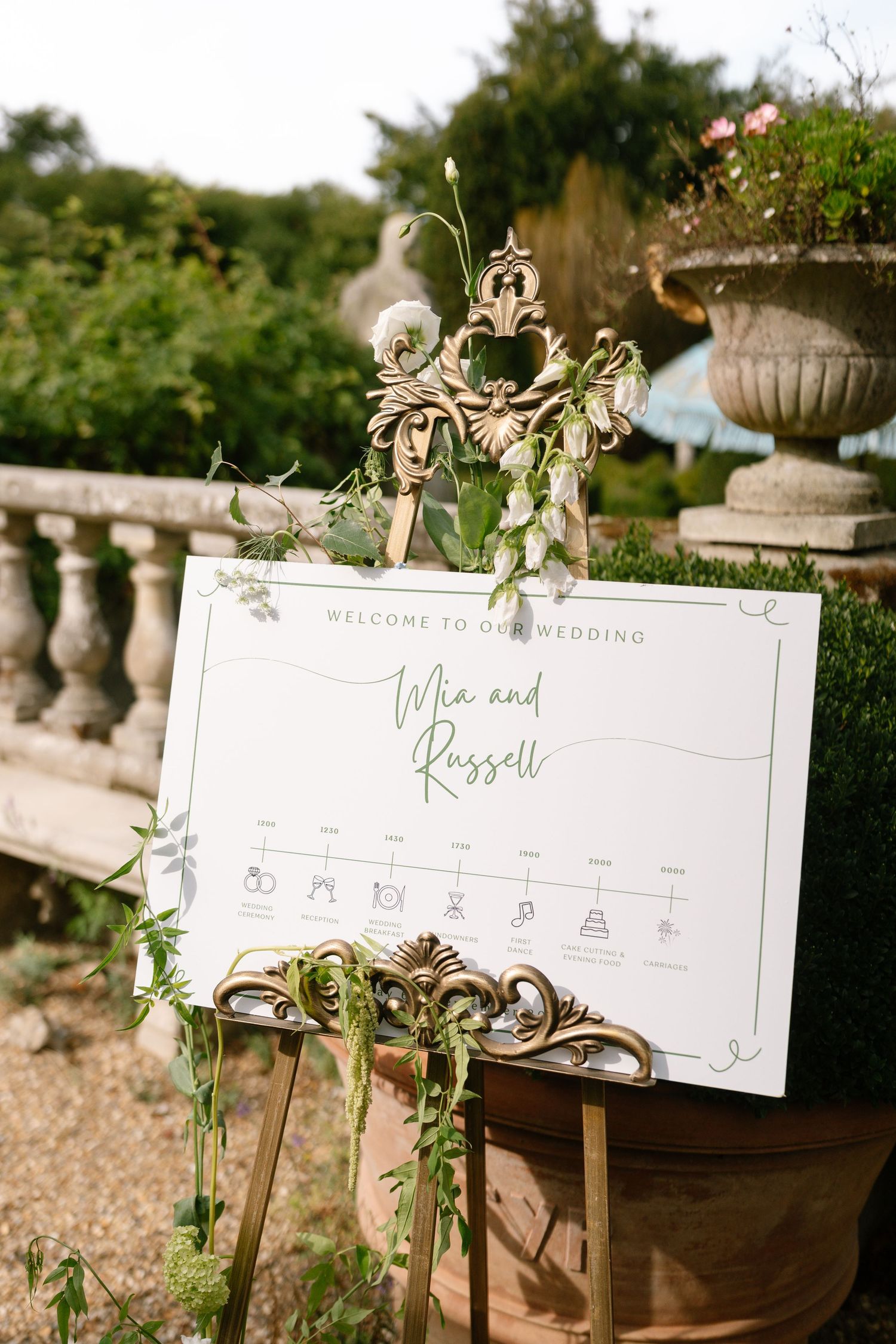 Wedding timeline and welcome signs decorated with greenery on wooden stands.