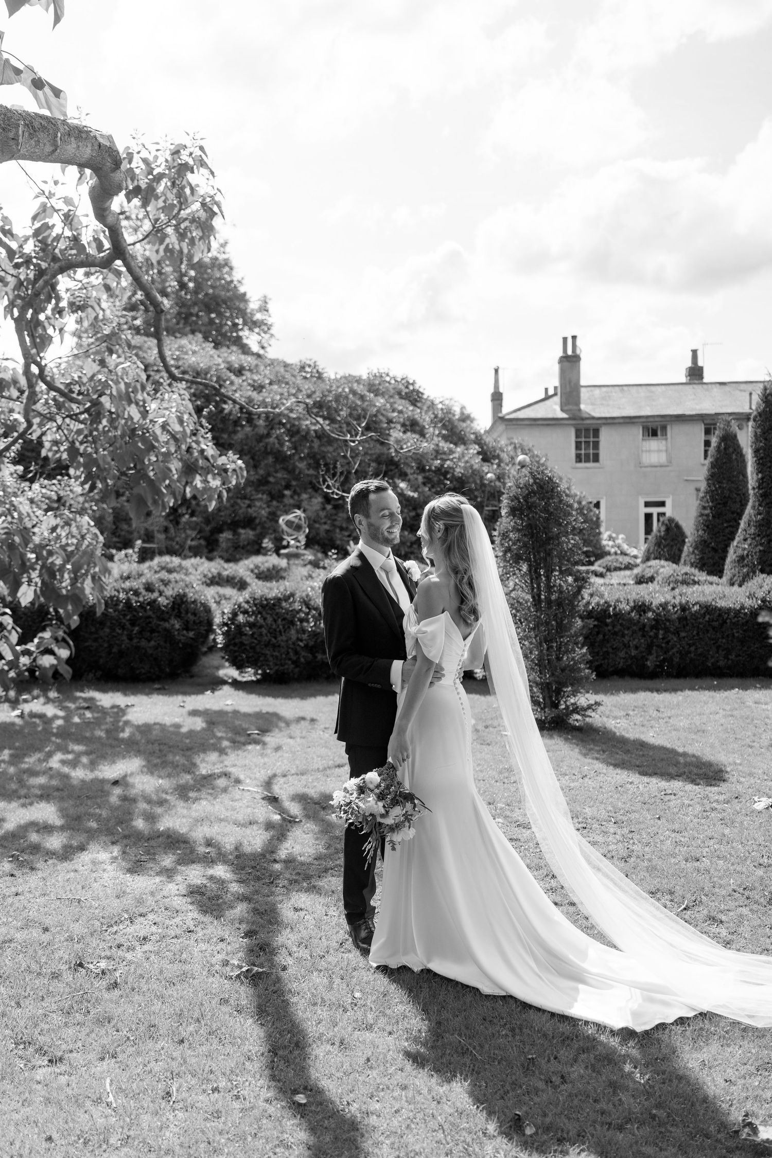 Black and white wedding photos captured in a scenic garden with a manor house backdrop.