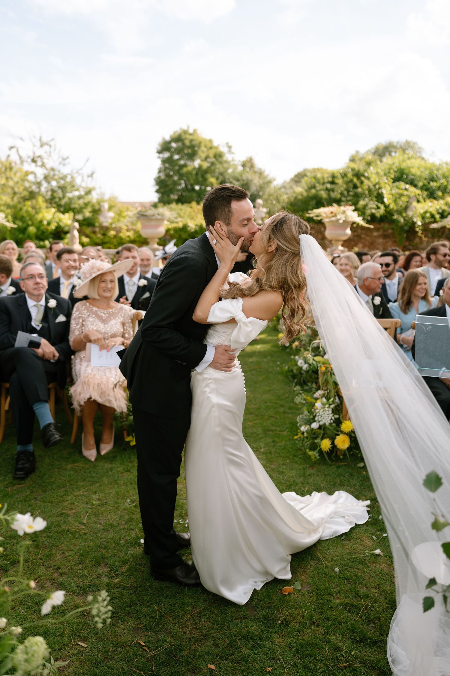 Bride and groom share passionate kiss during outdoor ceremony as guests look on.