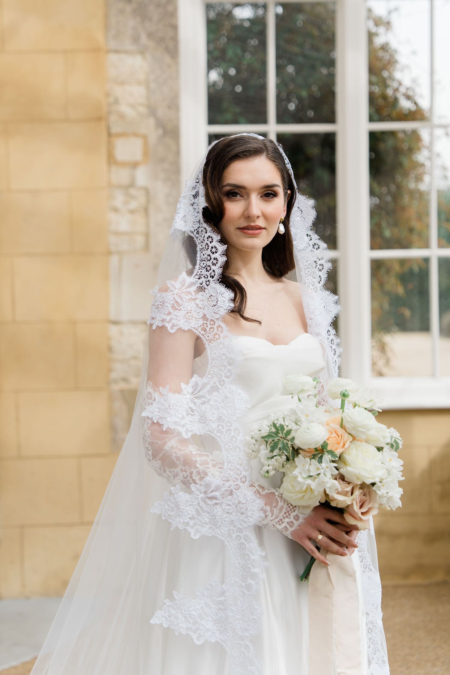 Elegant bride in lace veil holding white bouquet against sandstone building.