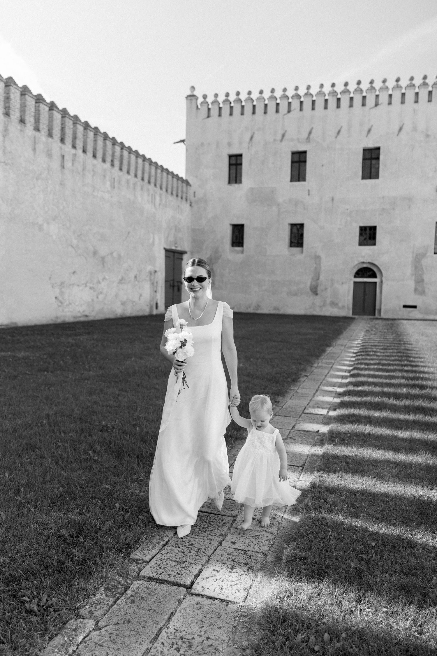 Black and white wedding photo showing figures walking along stone path by castle wall.