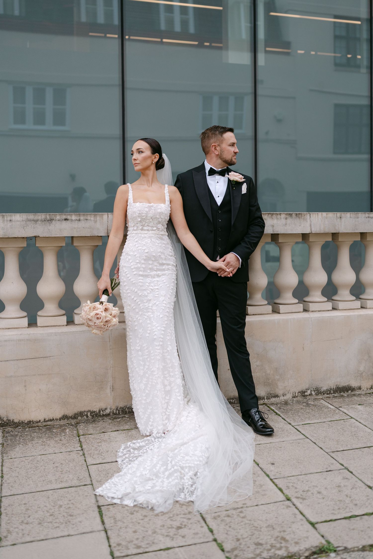 Wedding couple poses against glass building with stone balustrade in elegant series.