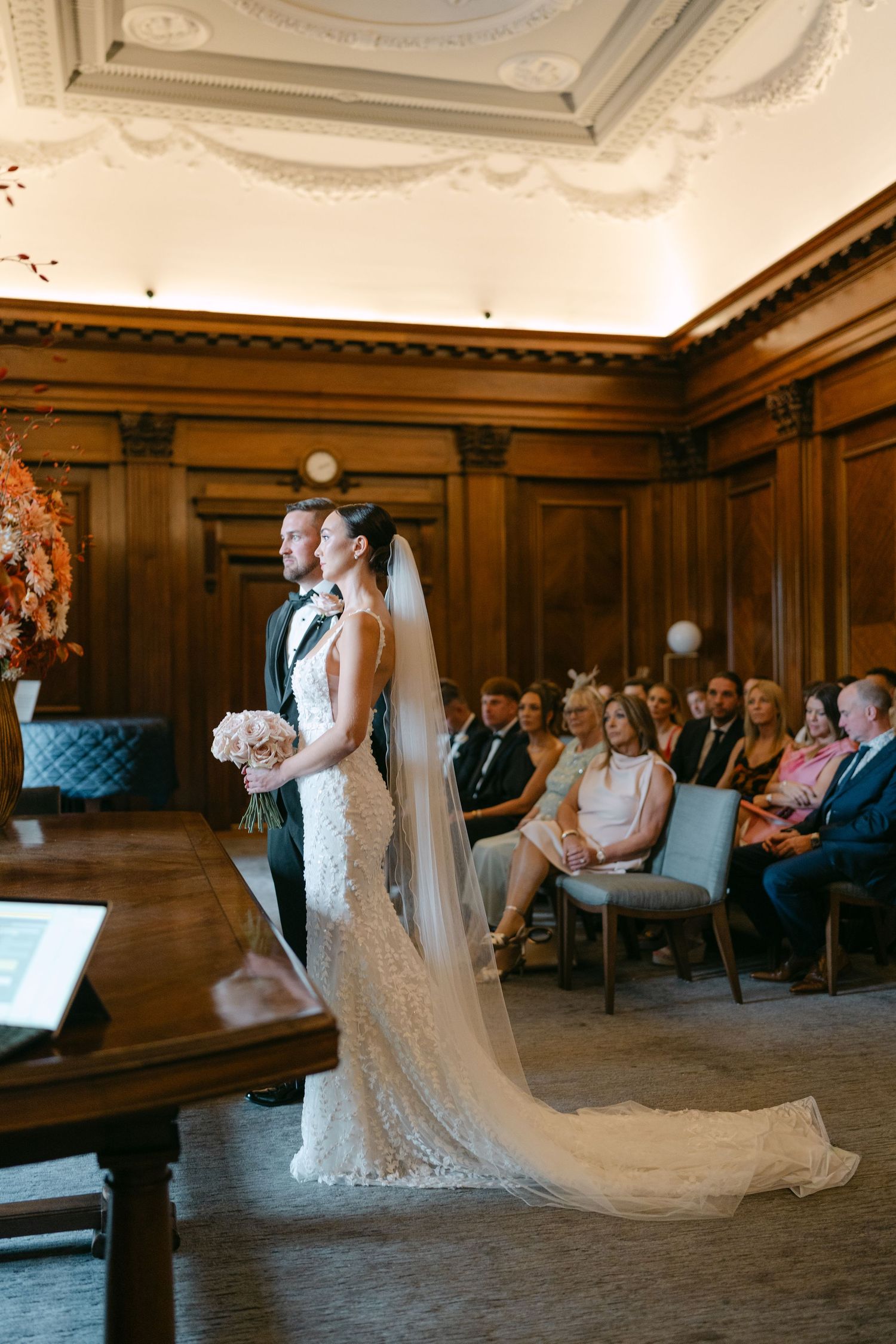 An elegant wedding ceremony in a grand wood-paneled room with ornate ceiling details.