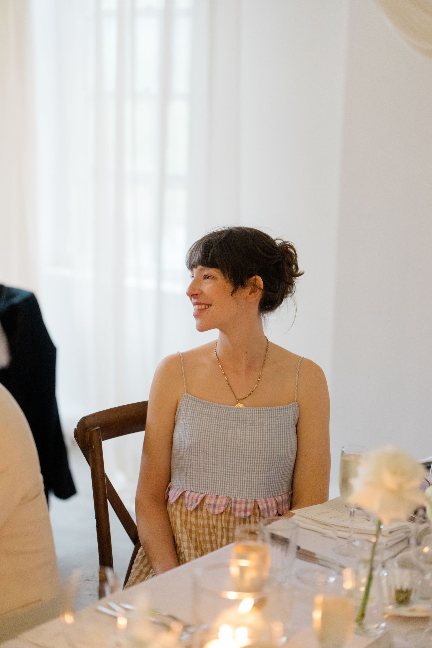 Smiling dark-haired female wedding guest in gray top seated at candlelit reception dinner table.