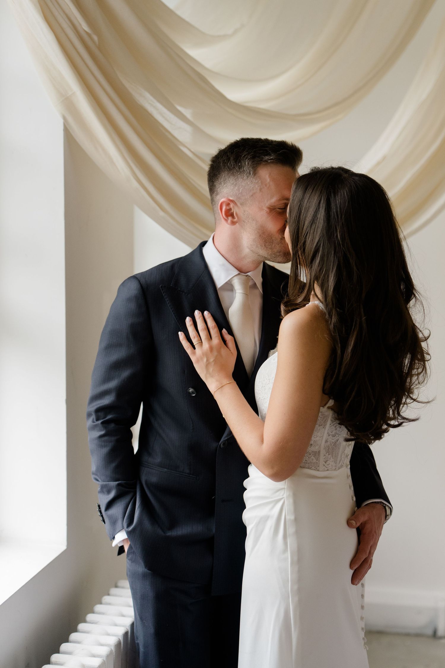 Bride and groom sharing a romantic kiss in front of elegant draped cream curtains.