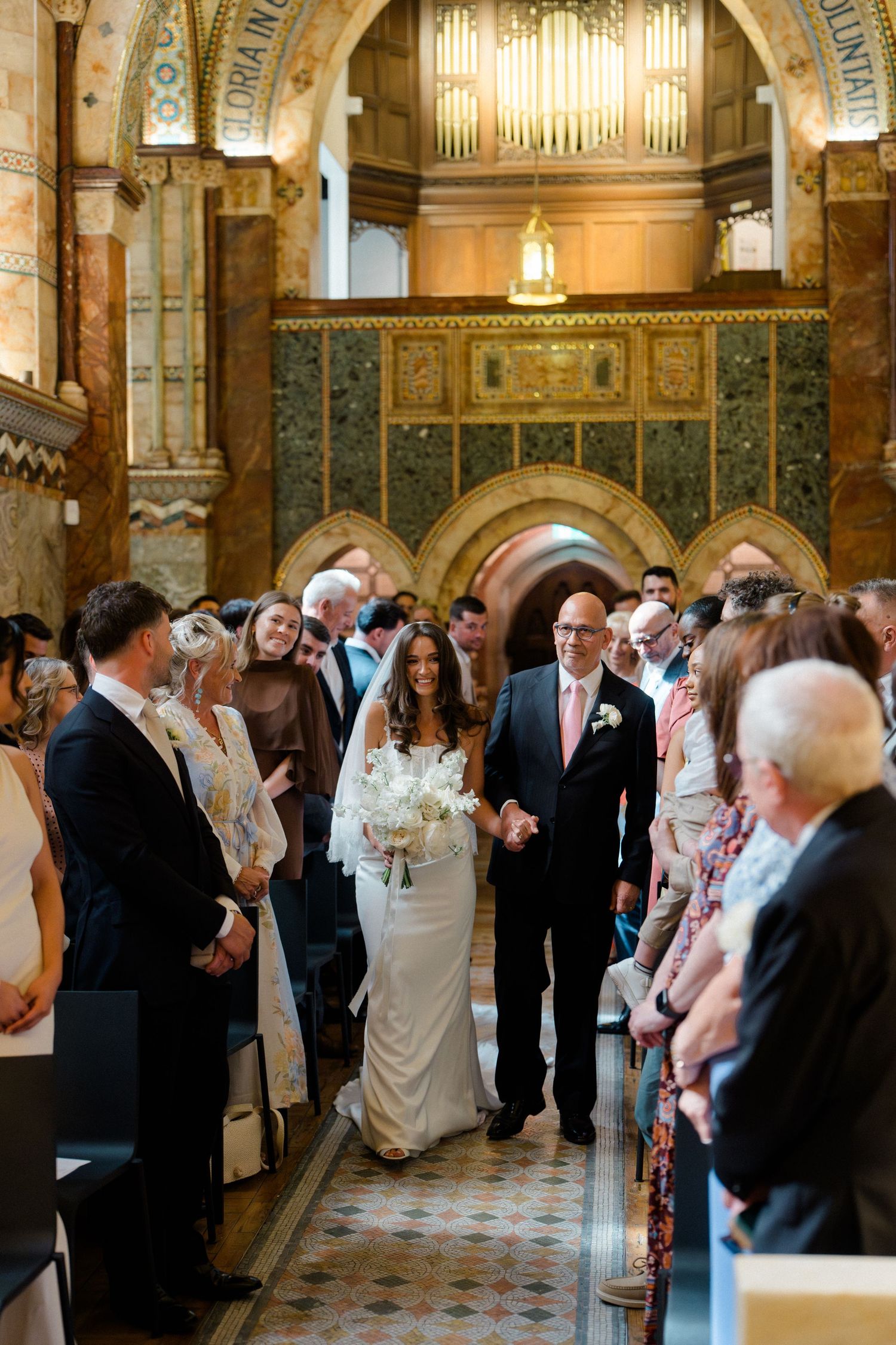 Bride walking down the aisle with her father in an ornate Byzantine-style church filled with wedding guests.