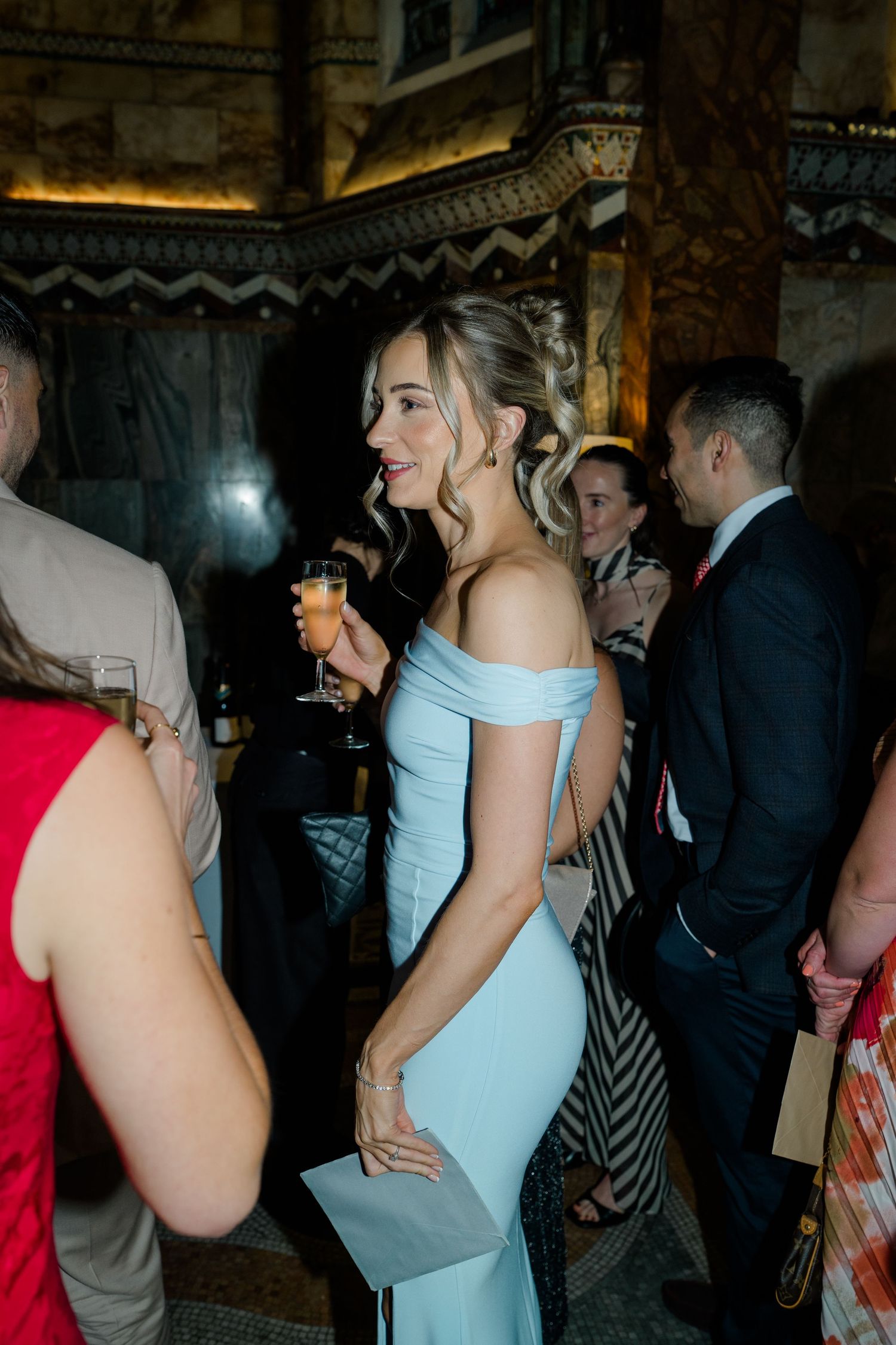 Bridesmaid in light blue off-shoulder gown holds champagne glass at elegant wedding reception venue.
