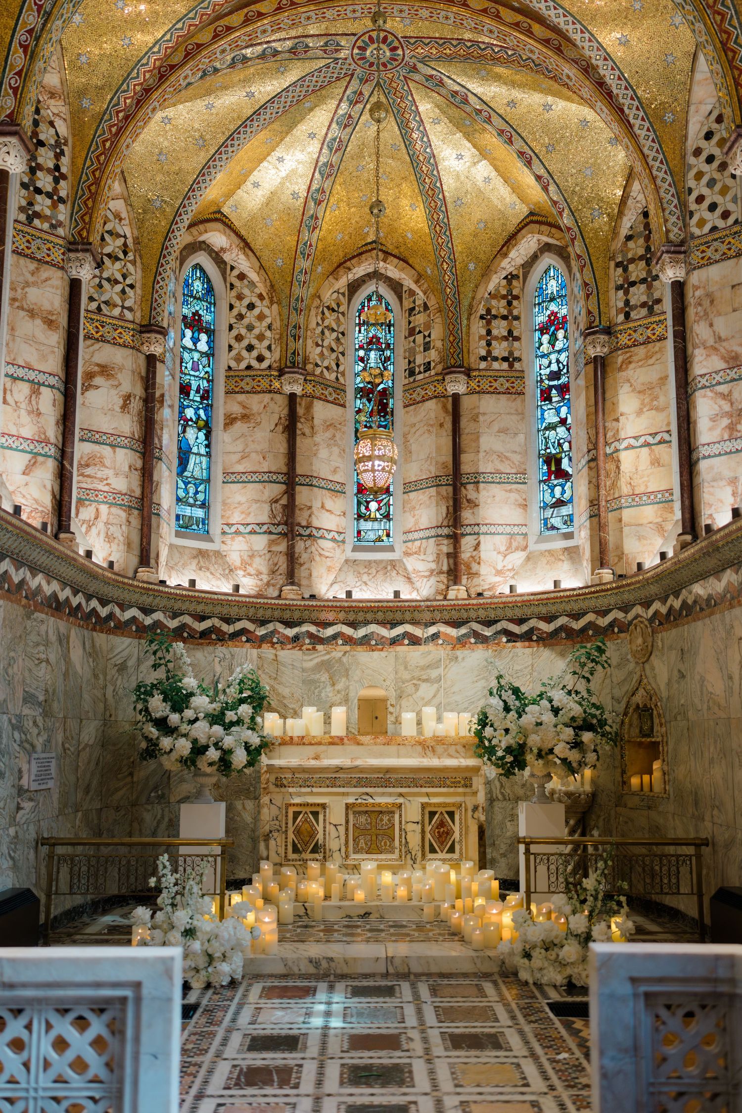 Ornate chapel altar adorned with white floral arrangements and candles beneath a stunning golden mosaic vaulted ceiling.