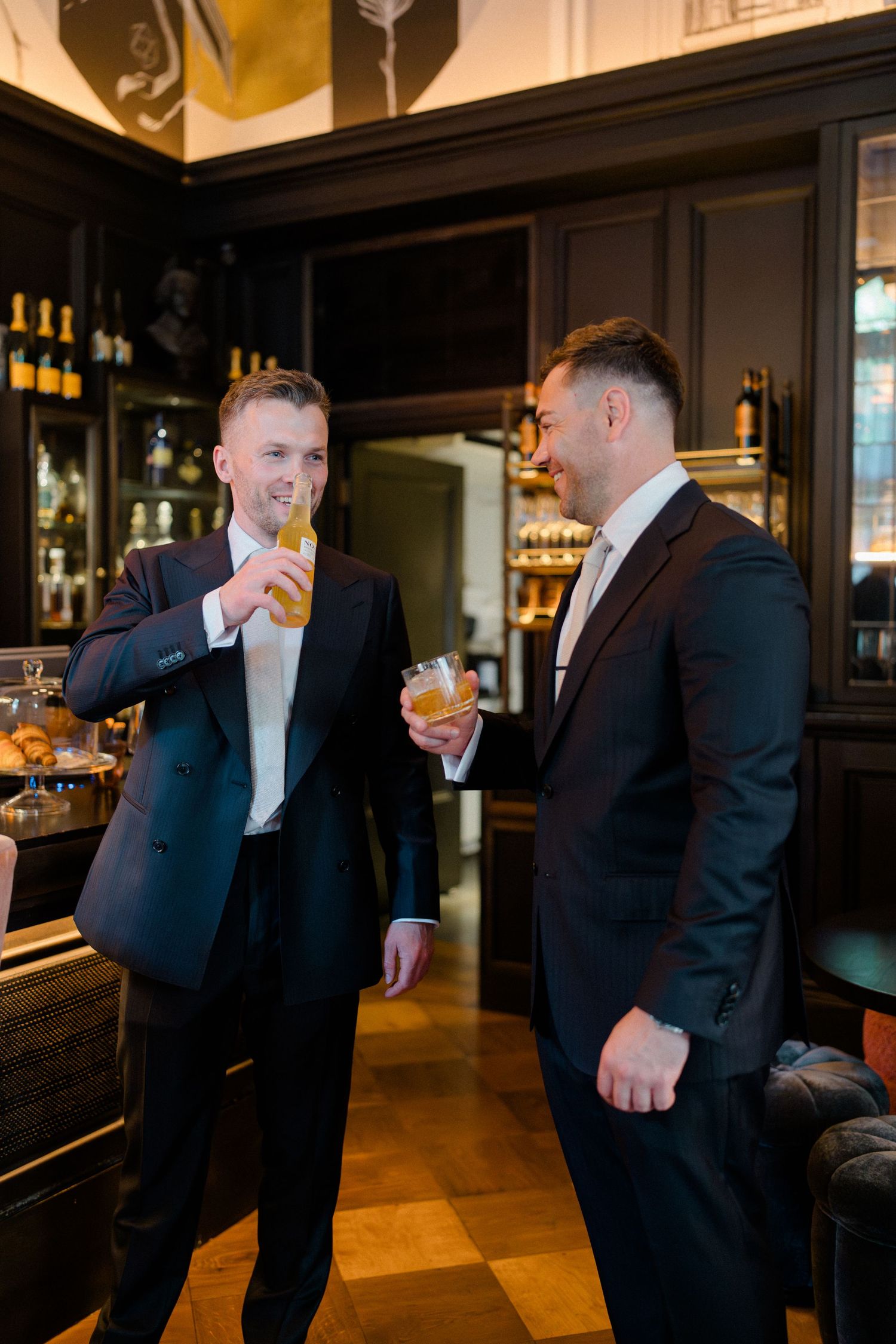 Two grooms in black suits share drinks and laugh together in an elegant dark wood bar setting.