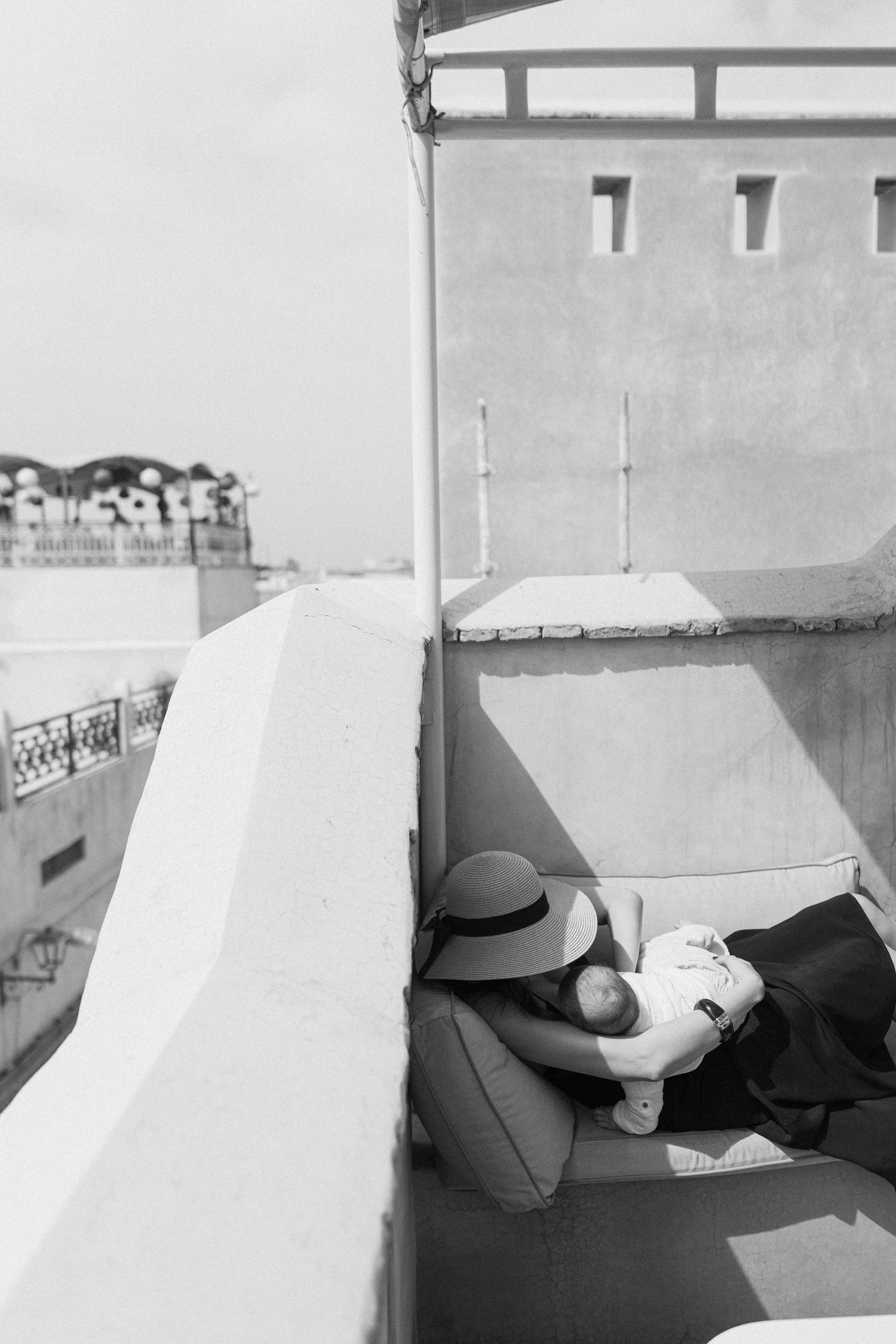 Black and white photo of a woman in a hat resting on a balcony chair overlooking a European cityscape.