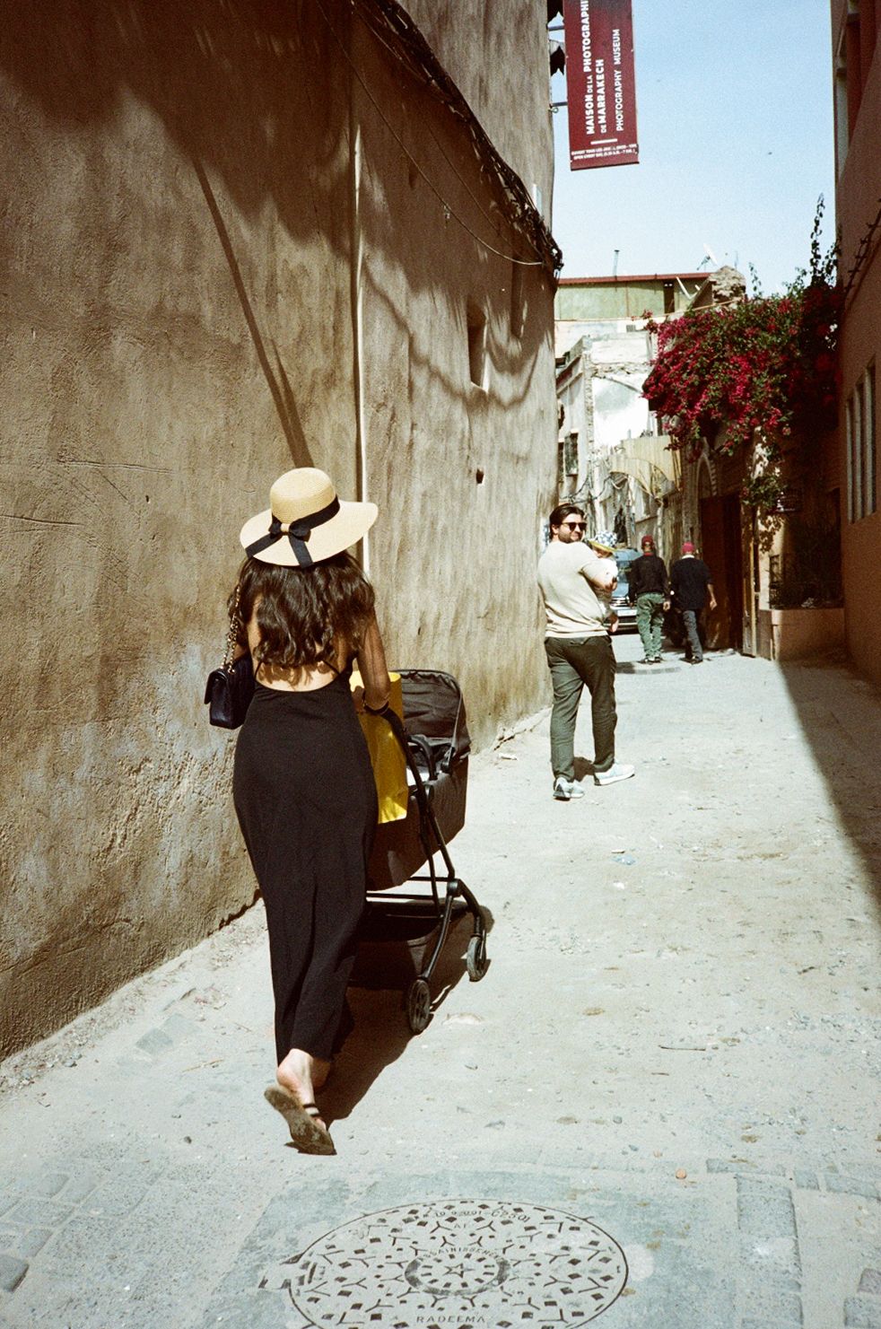 Woman in black dress and straw hat pushes stroller through narrow sunlit alley in historic city.