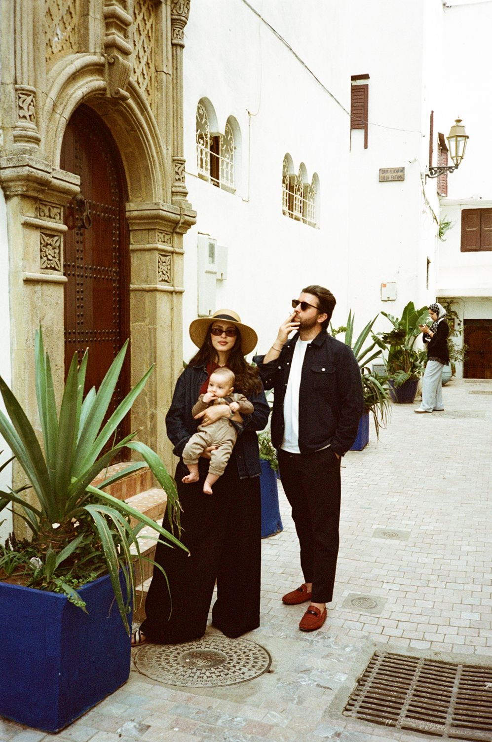 A stylish couple with a baby poses in a charming Mediterranean alley near an ornate wooden door.