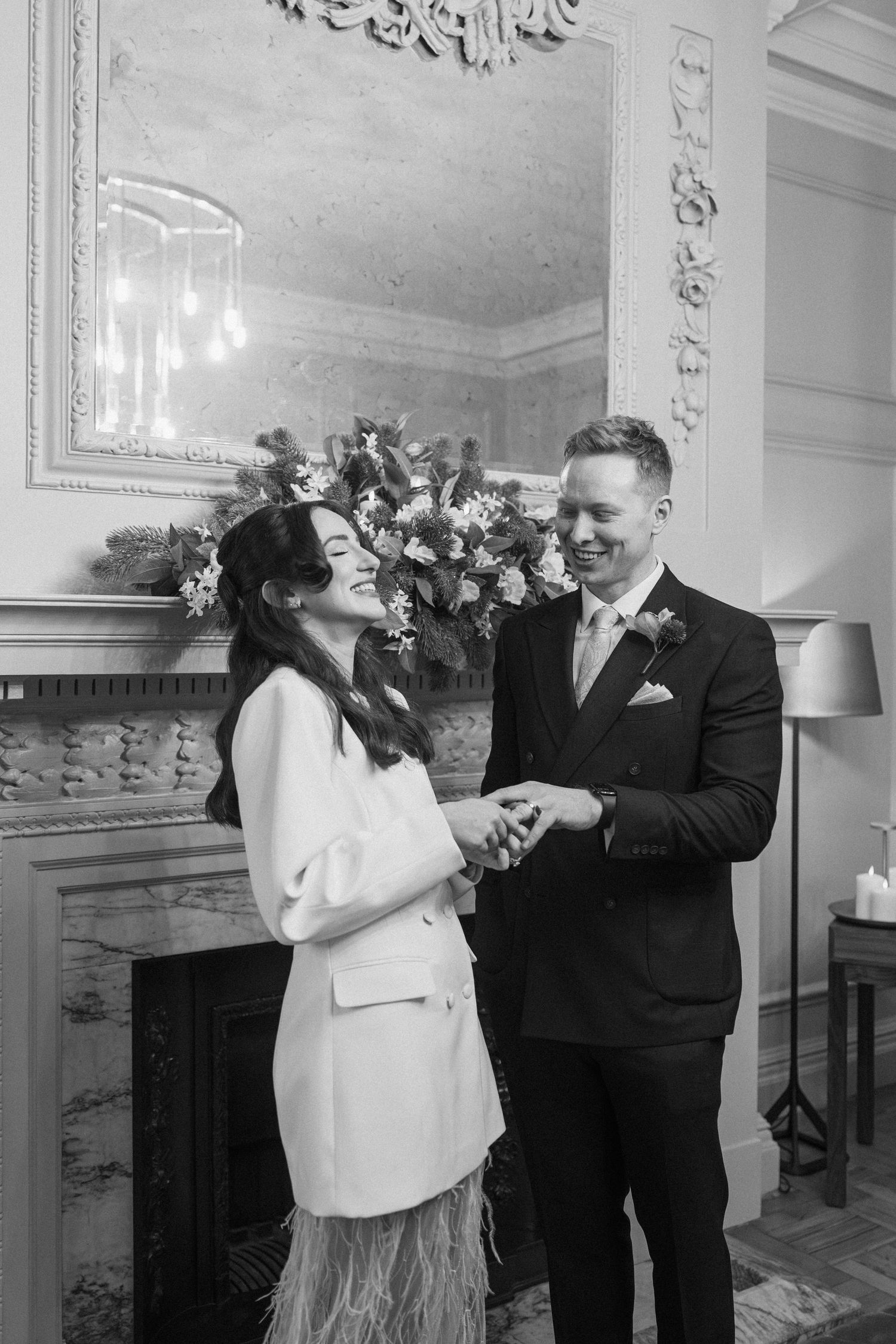 Black and white photo of laughing bride and groom exchanging rings by ornate fireplace.