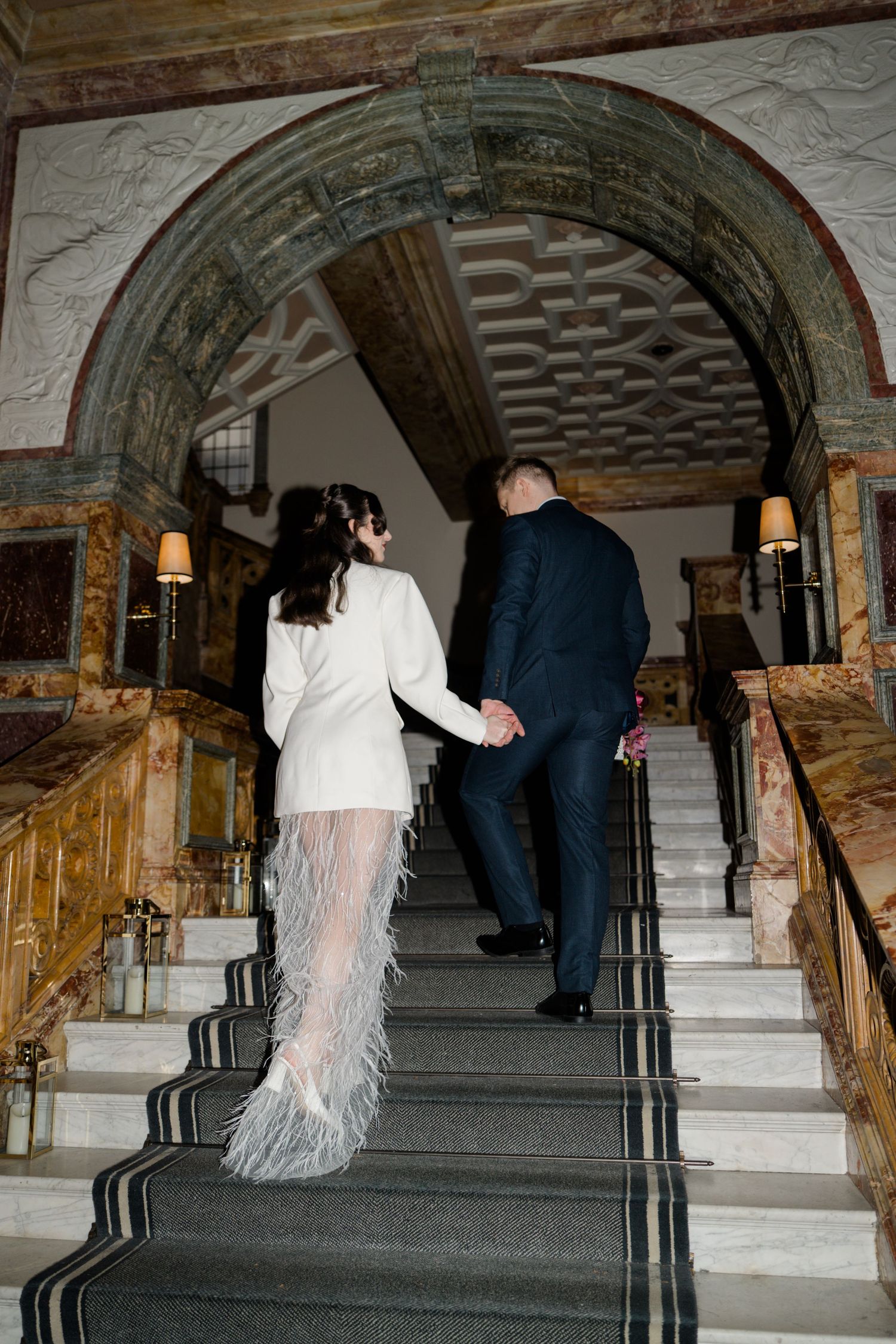 Bride in white feather mini dress and groom in navy suit ascending grand marble staircase.