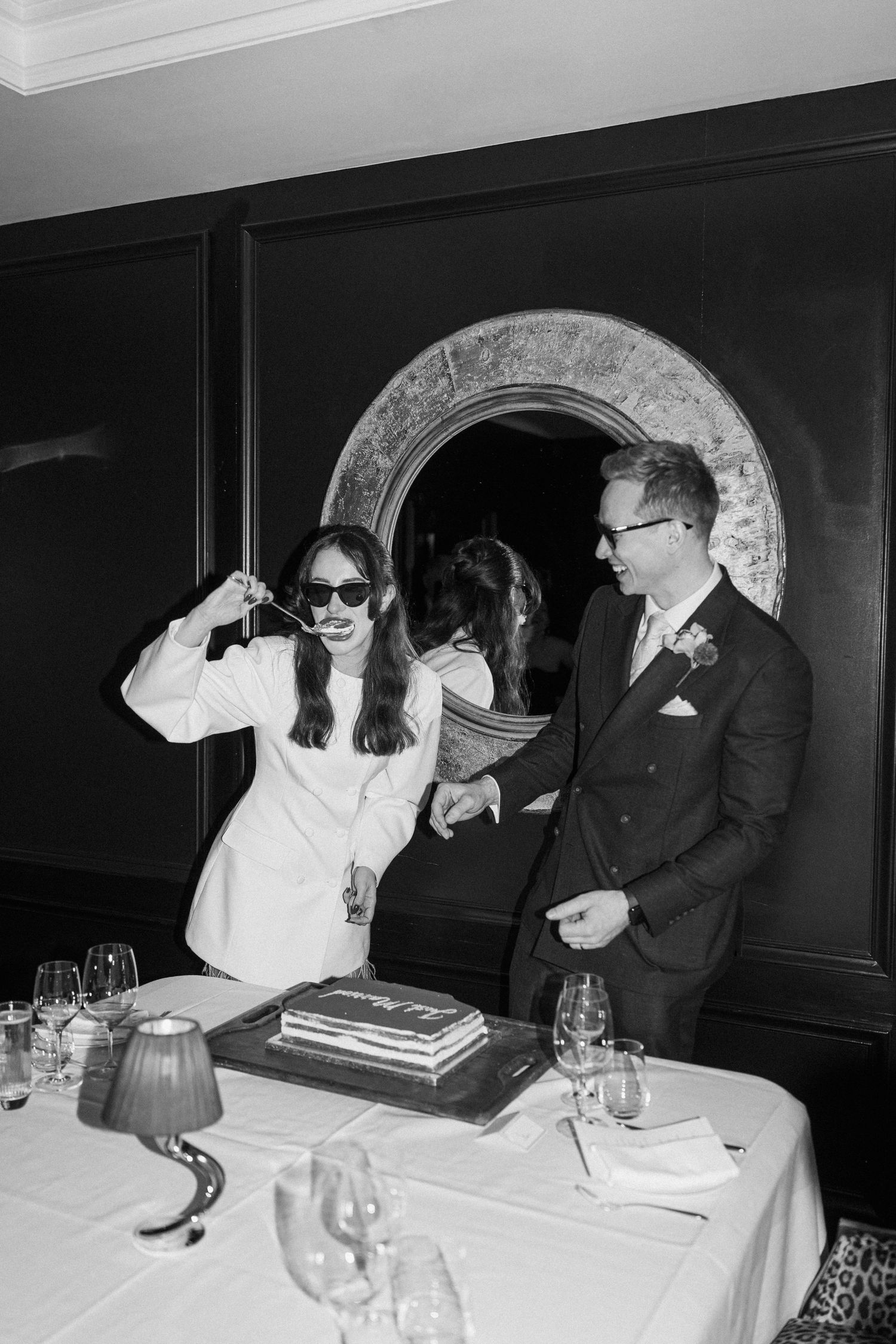 Bride in sunglasses and white blazer cutting wedding cake with groom in black and white.