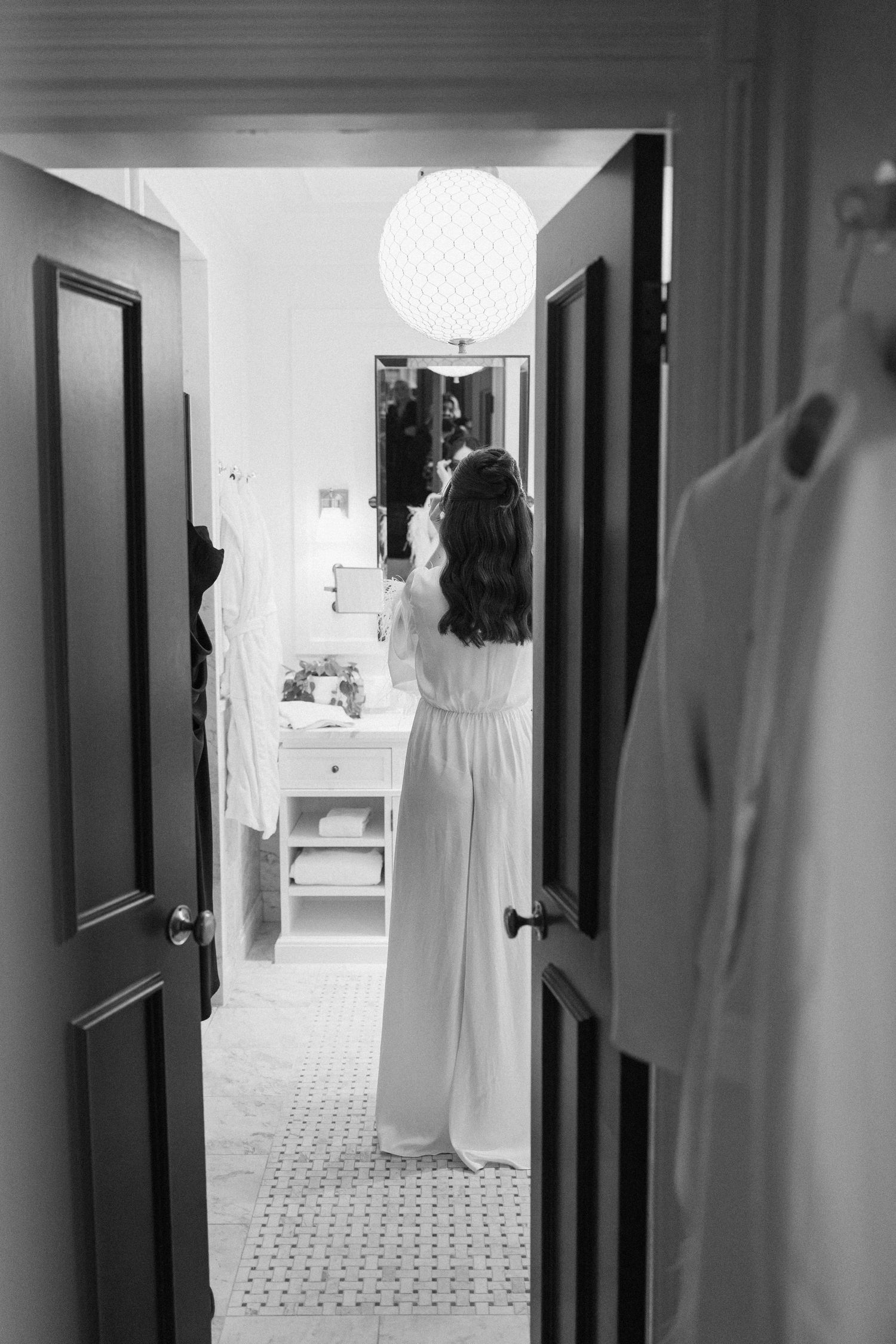 Bride in white dress preparing in bathroom mirror, black and white wedding getting ready photo.