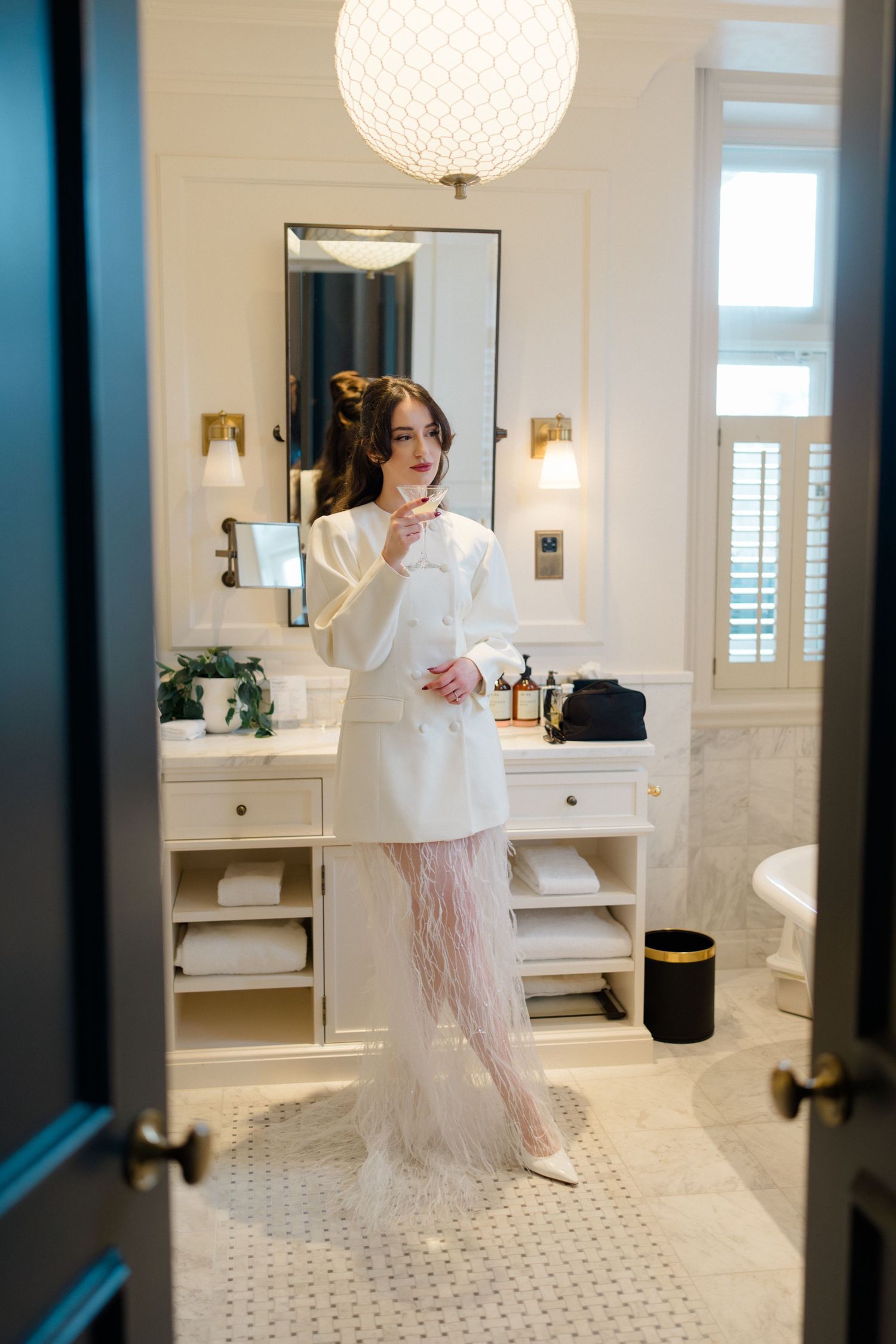 Bride in white blazer mini dress with feather skirt applying makeup in elegant marble bathroom.