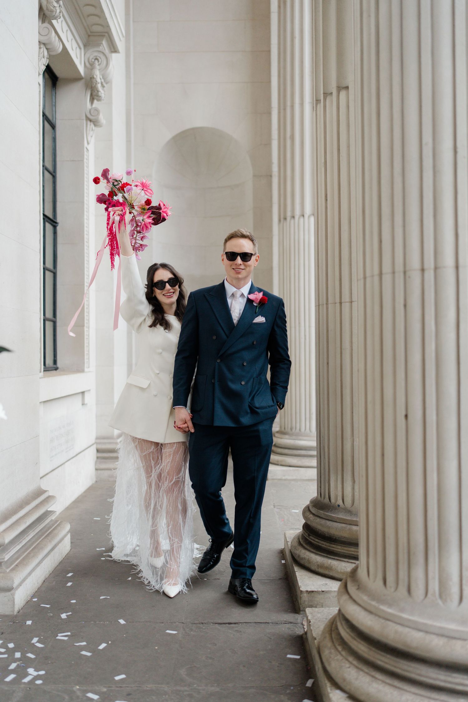 Bride and groom holding pink bouquet walking between grand columns after London registry wedding.