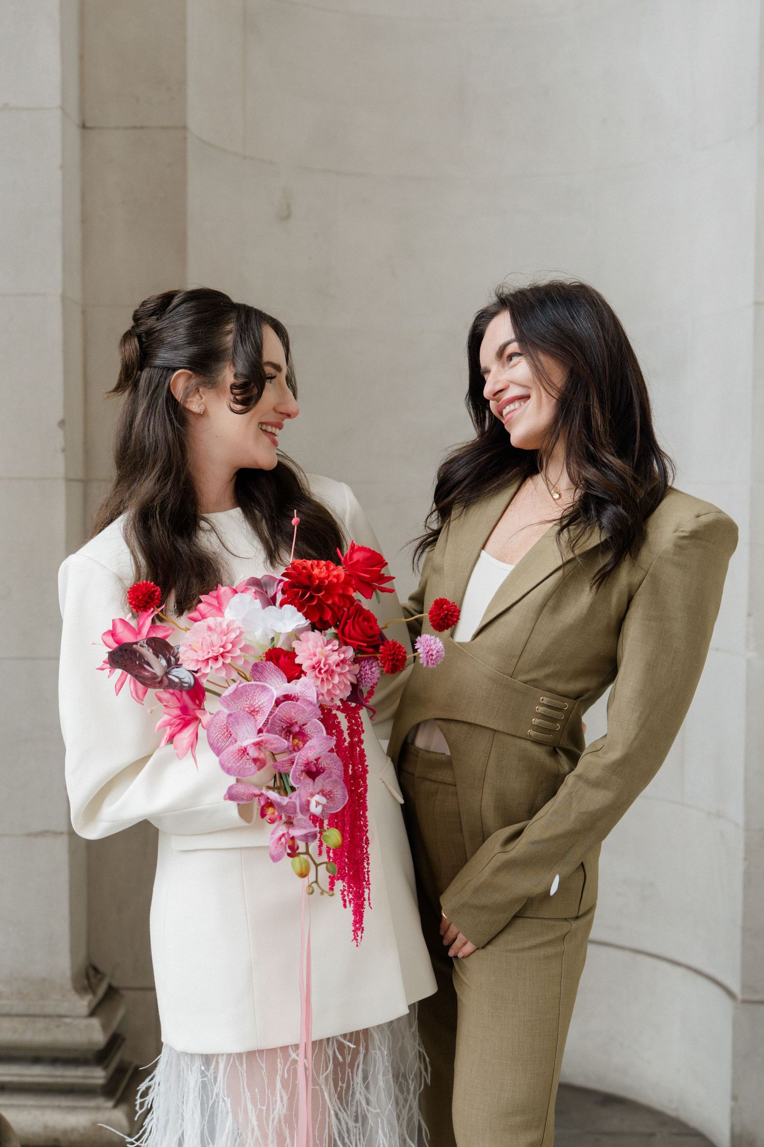 Two brides smile at each other, one in white feather dress holding pink bouquet, other in olive suit.