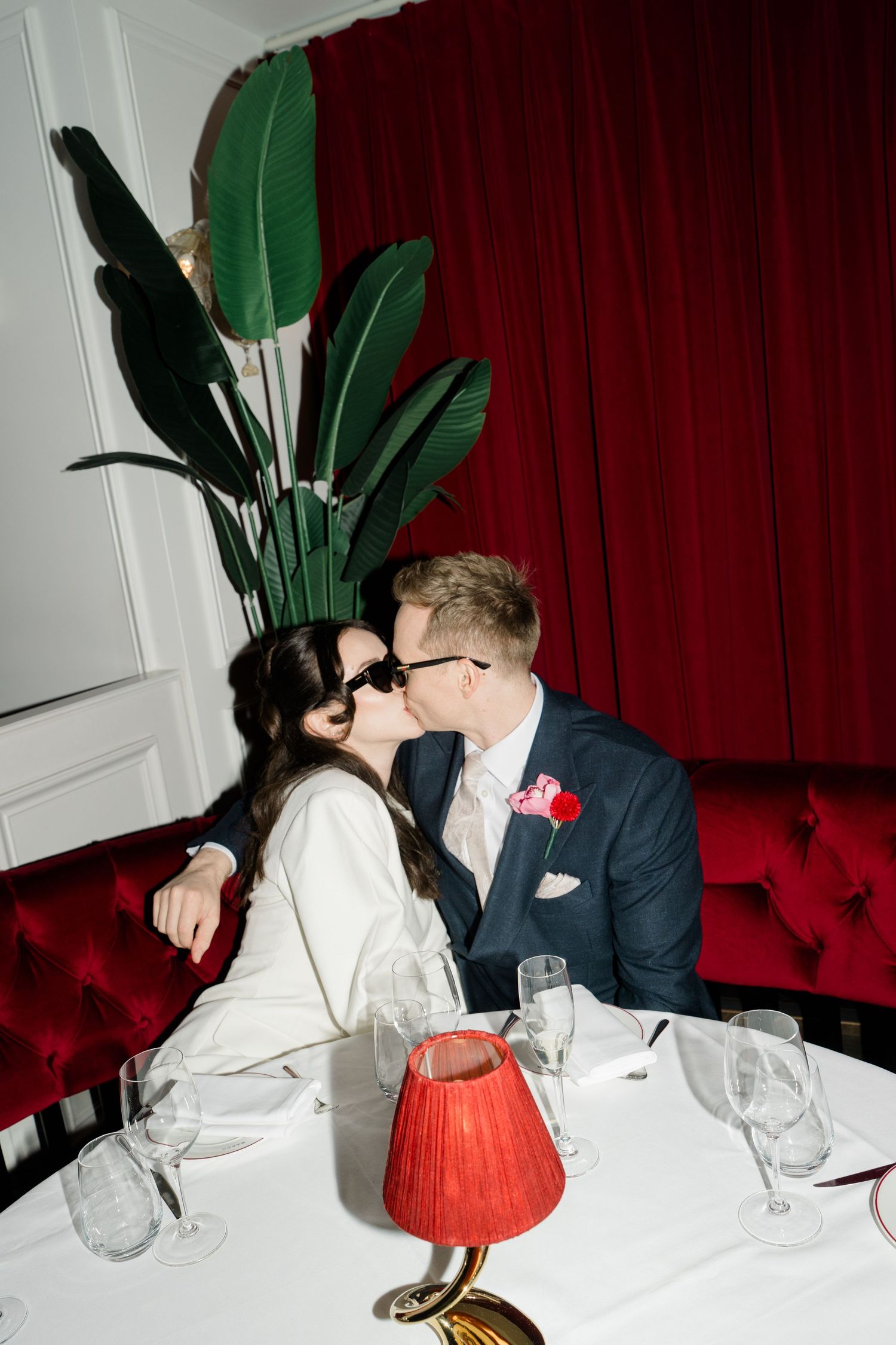 Newlyweds kiss in red velvet booth at elegant restaurant table with crystal glasses and red lamp.