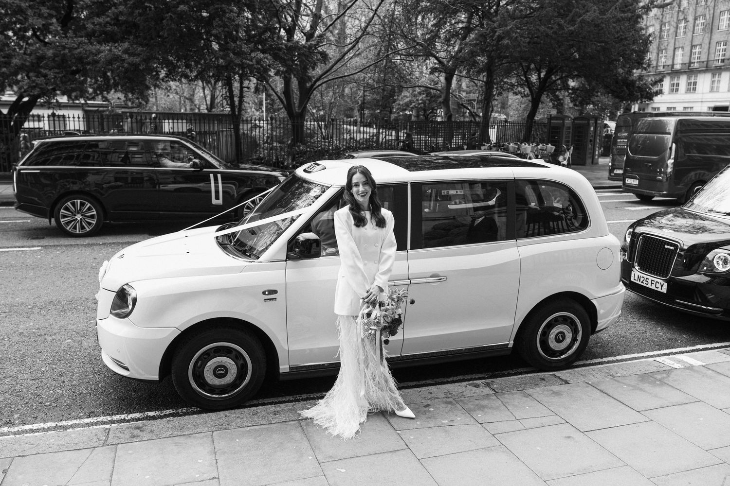 Bride in long white gown poses beside a decorated white London taxi cab on a city street.