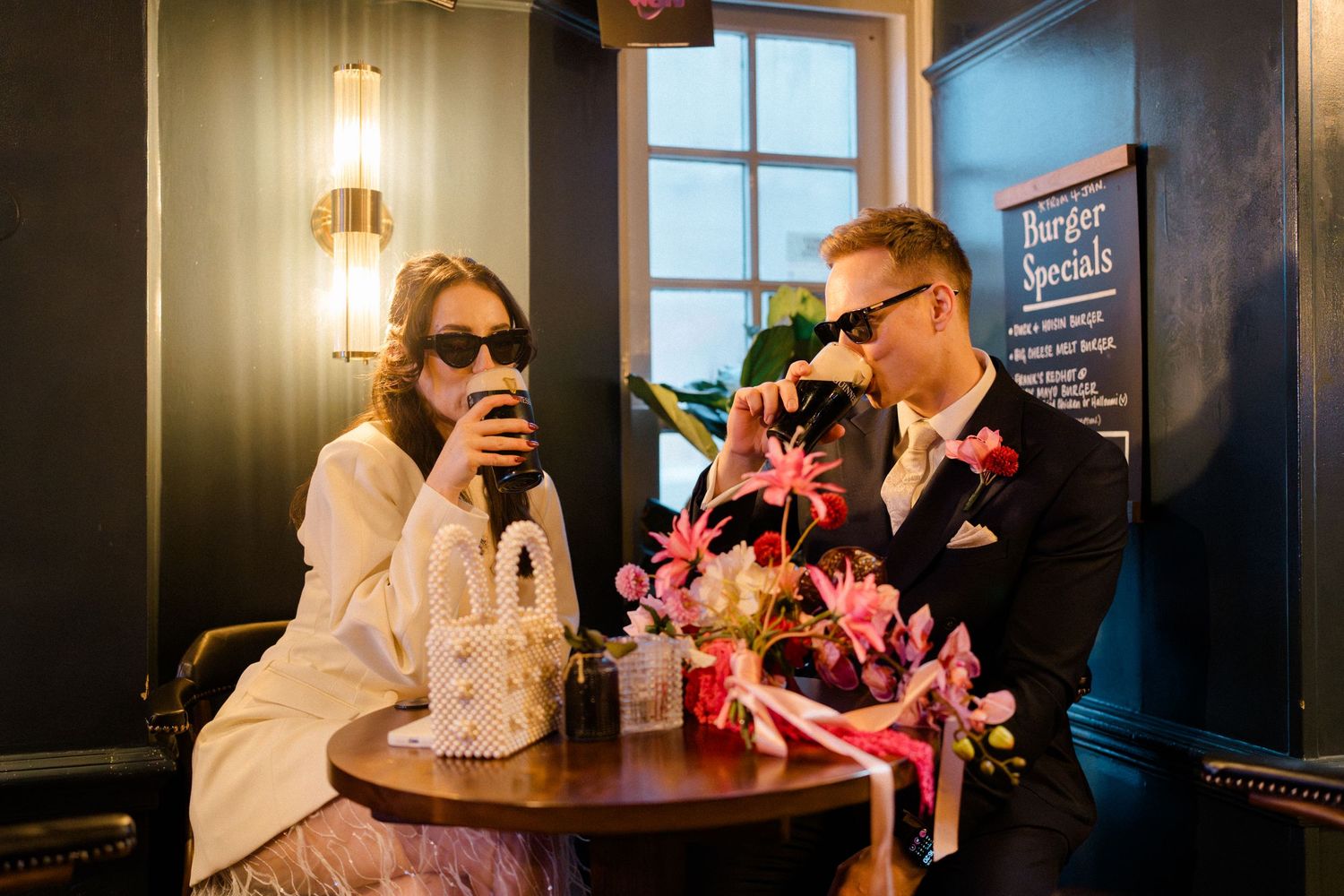 Newlyweds in sunglasses share drinks at dark pub table with floral centerpiece and burger specials menu.