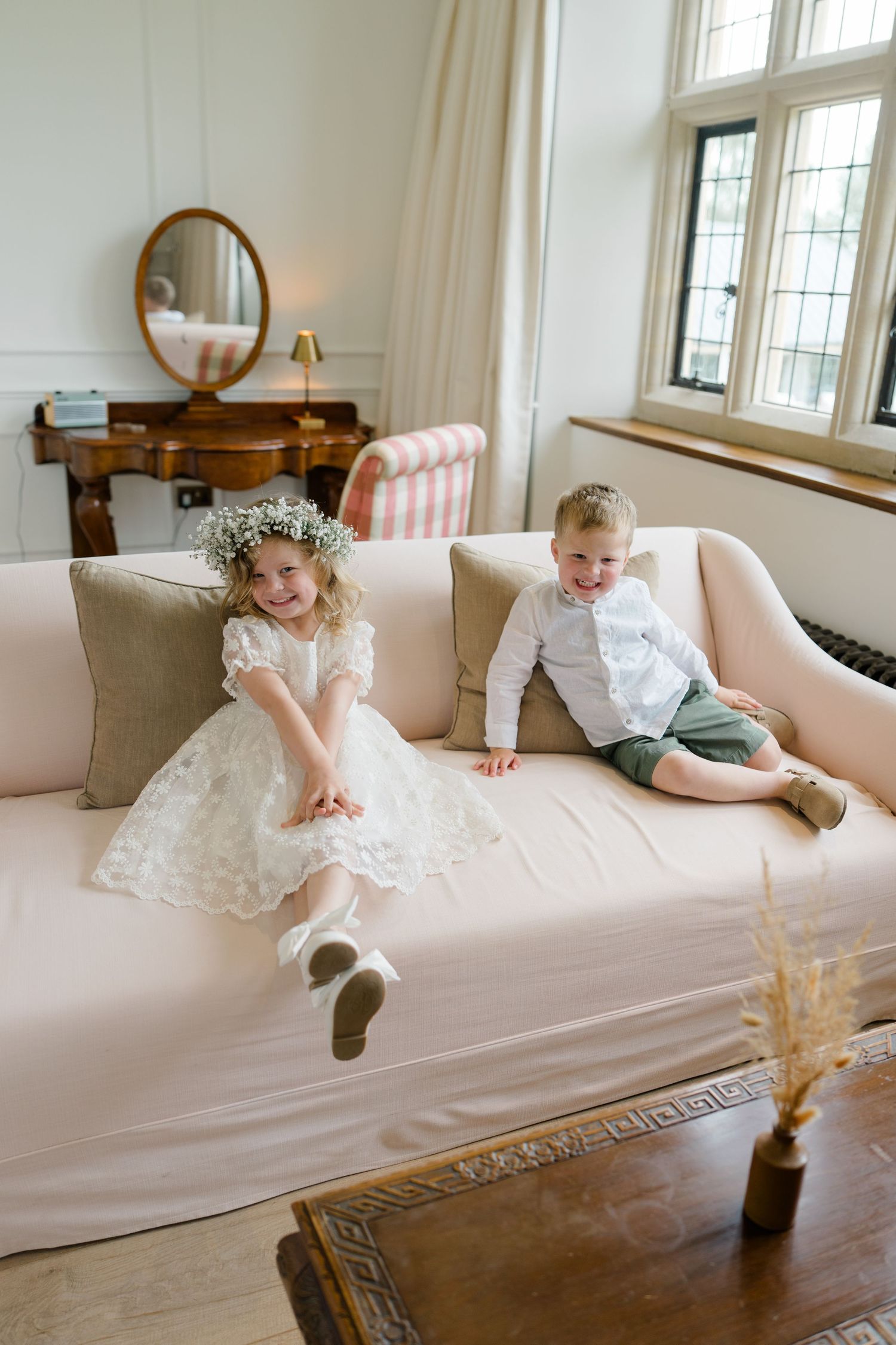 Two young children in formal wedding attire sit on a pink sofa, girl in white lace dress with flower crown, boy in white shirt.