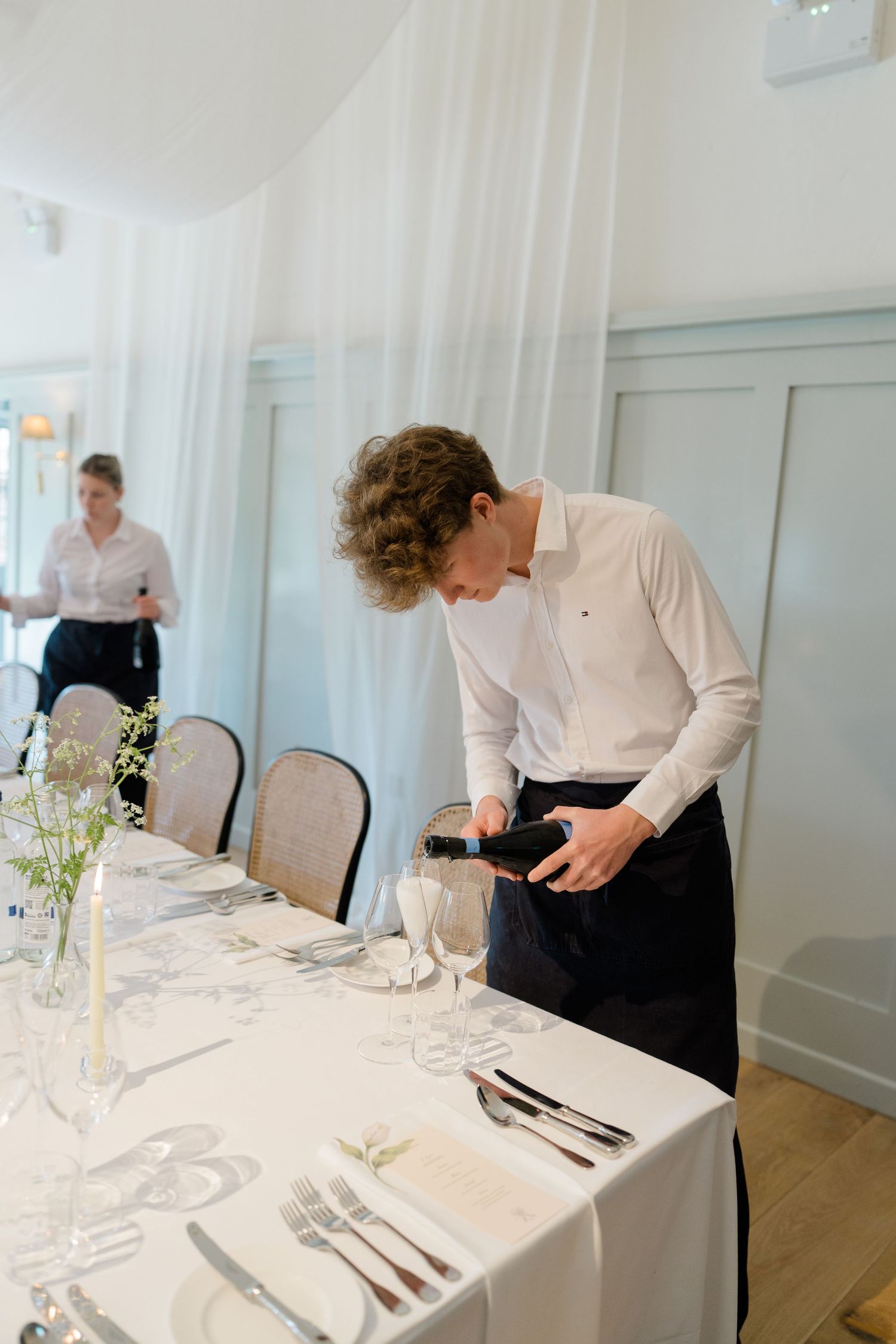 A young waiter in a white shirt pours wine at an elegantly set wedding reception table with white linens.