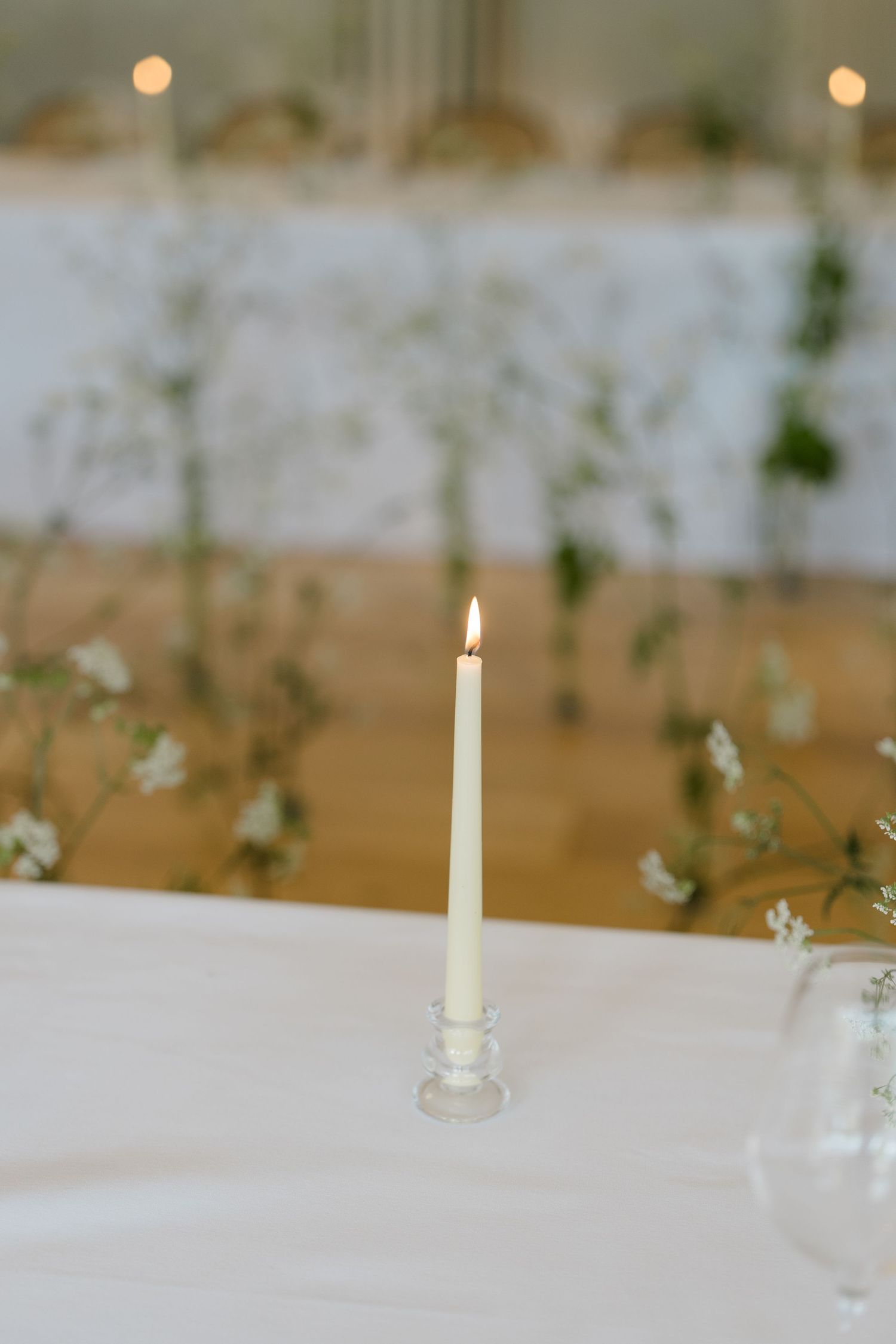 Single white taper candle in glass holder on white linen tablecloth with greenery wedding centerpieces.