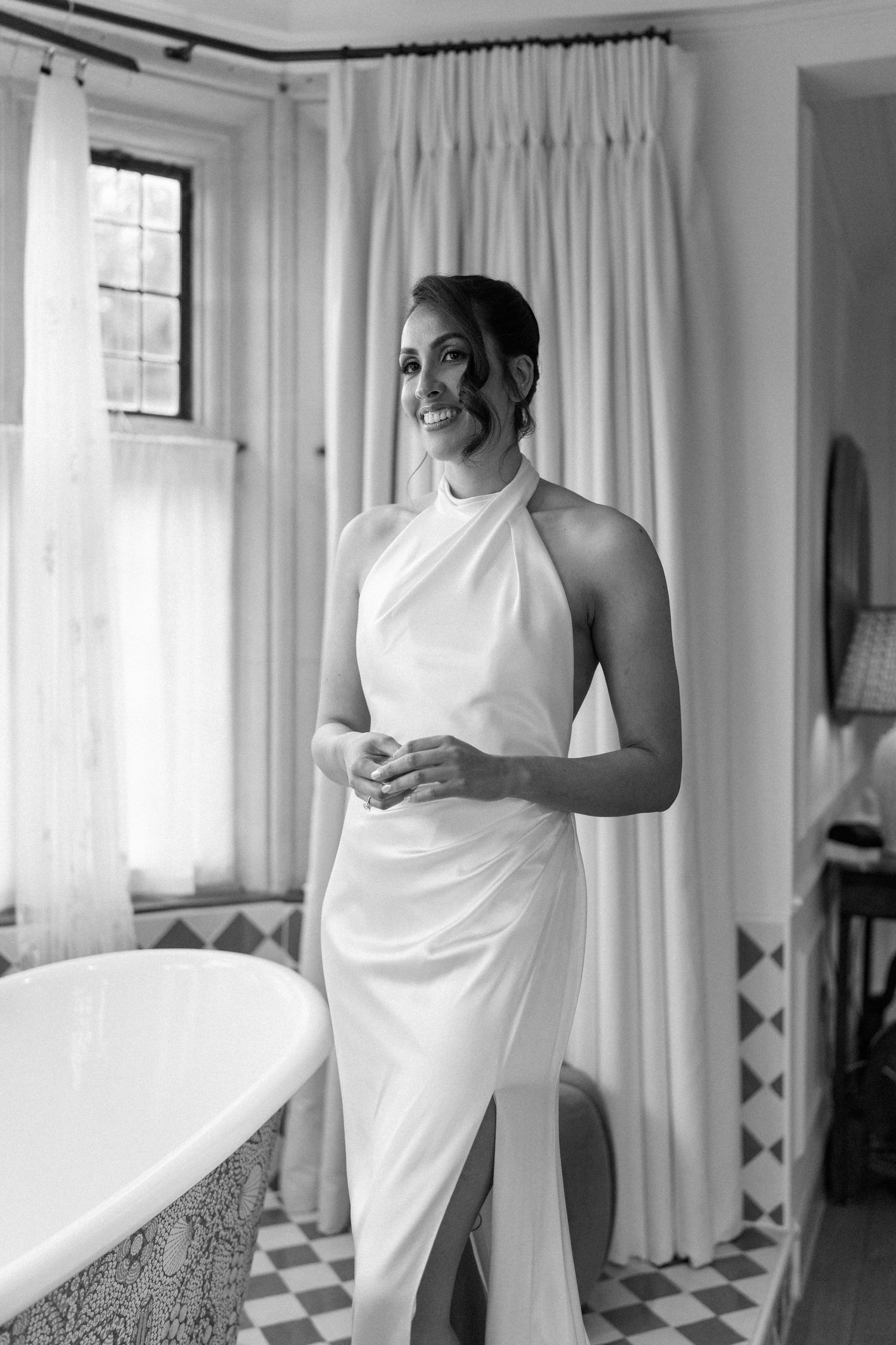 Bride in elegant halter neck wedding dress standing by a freestanding bathtub in a luxurious bathroom.