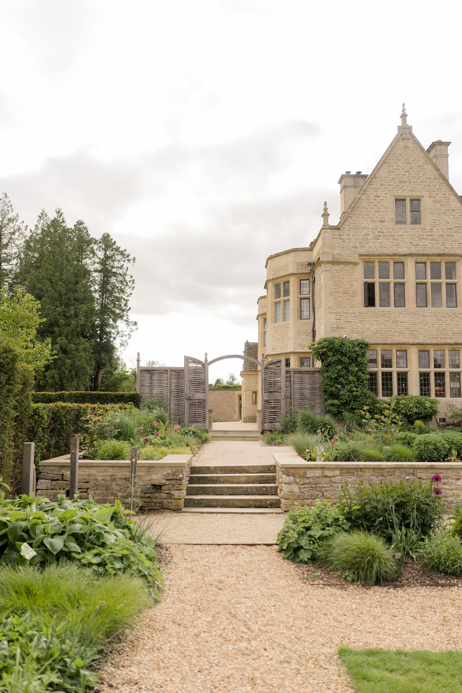 A grand Cotswold stone manor house with manicured gardens, stone steps, and a gravel pathway on an overcast day.