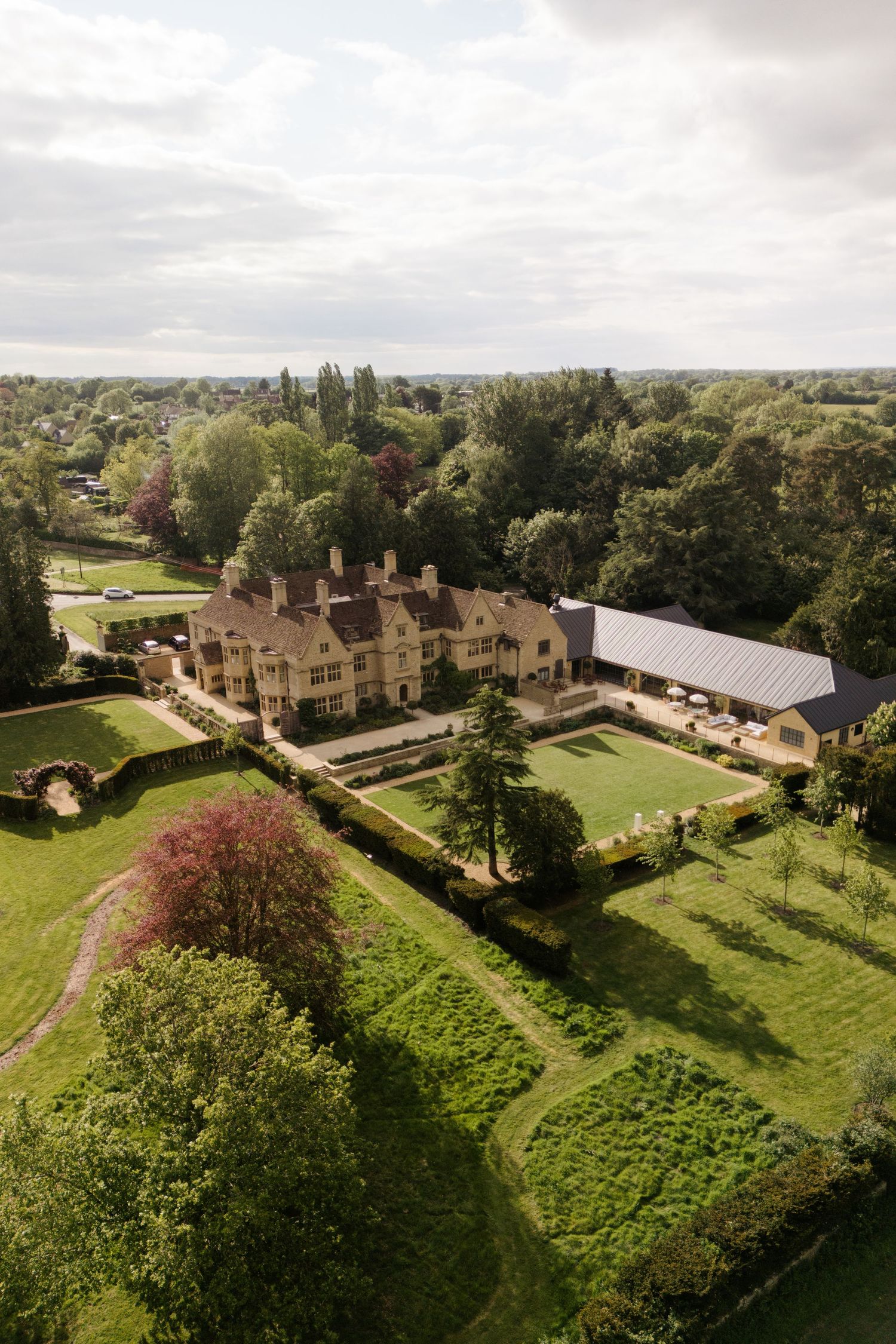 Aerial view of a grand English country manor house surrounded by manicured gardens and lush green trees.