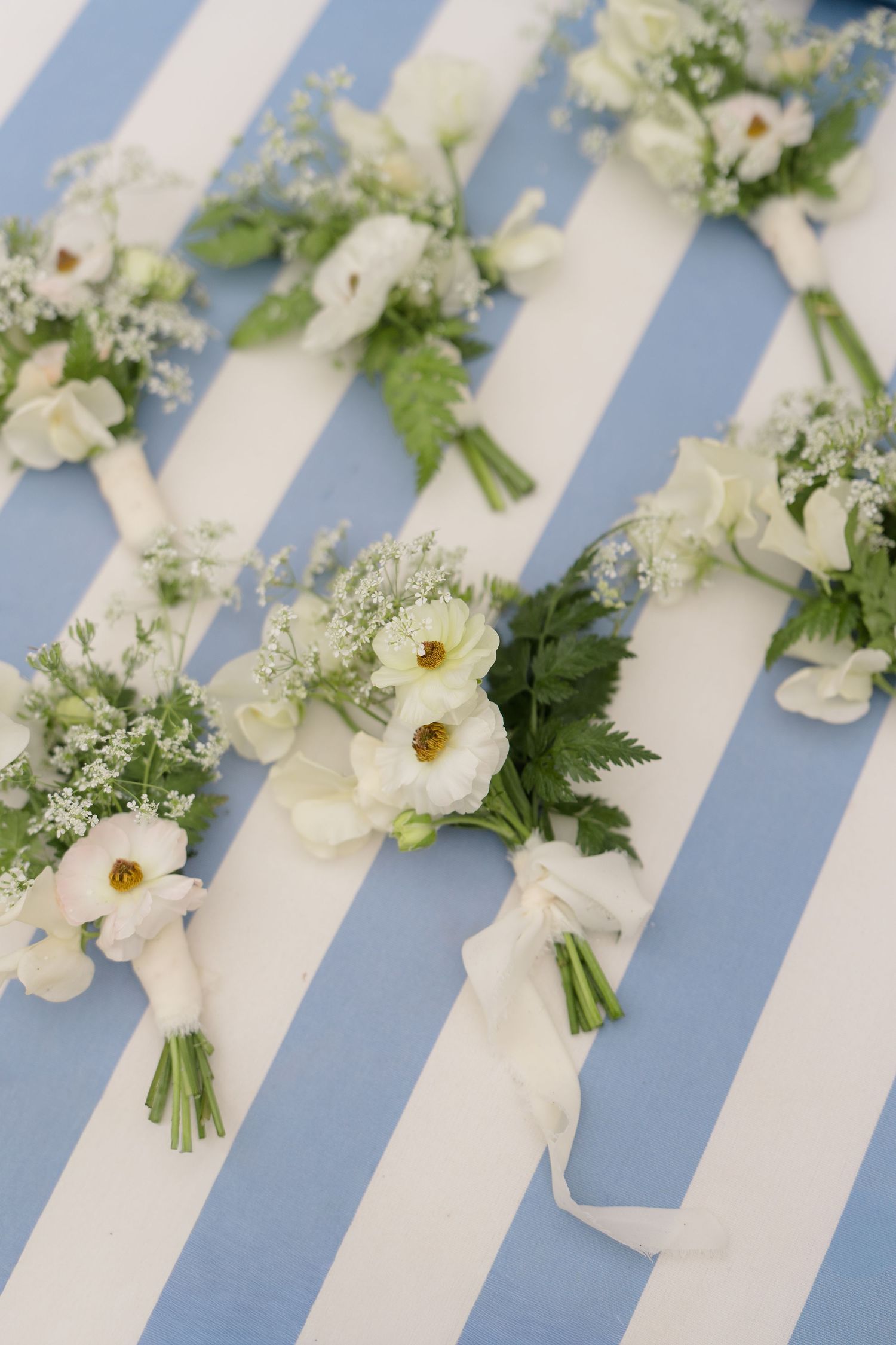 White wedding boutonnieres with anemones and greenery tied with ribbon on a blue and white striped surface.