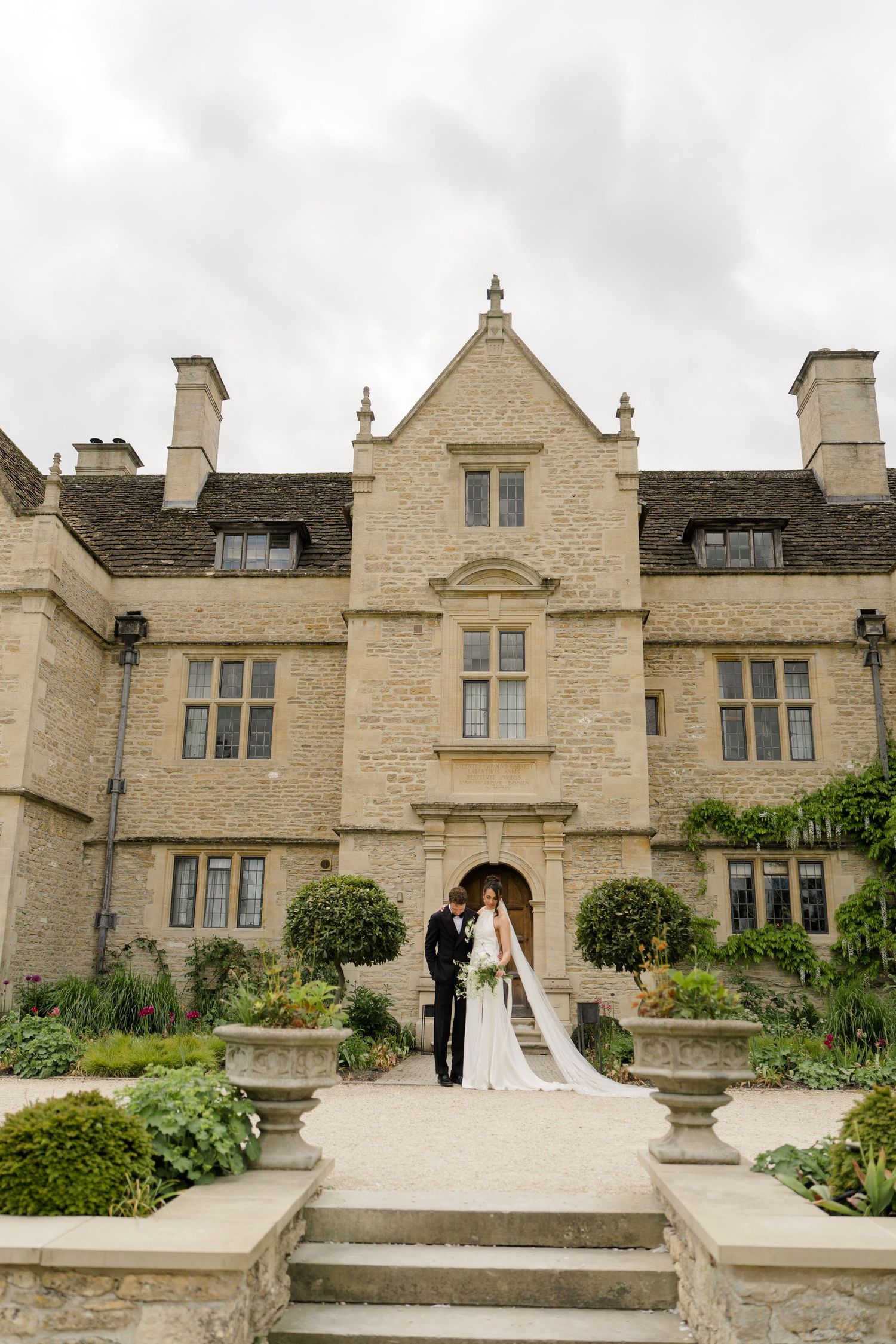 Bride and groom pose on steps of grand English stone manor house under dramatic cloudy sky.