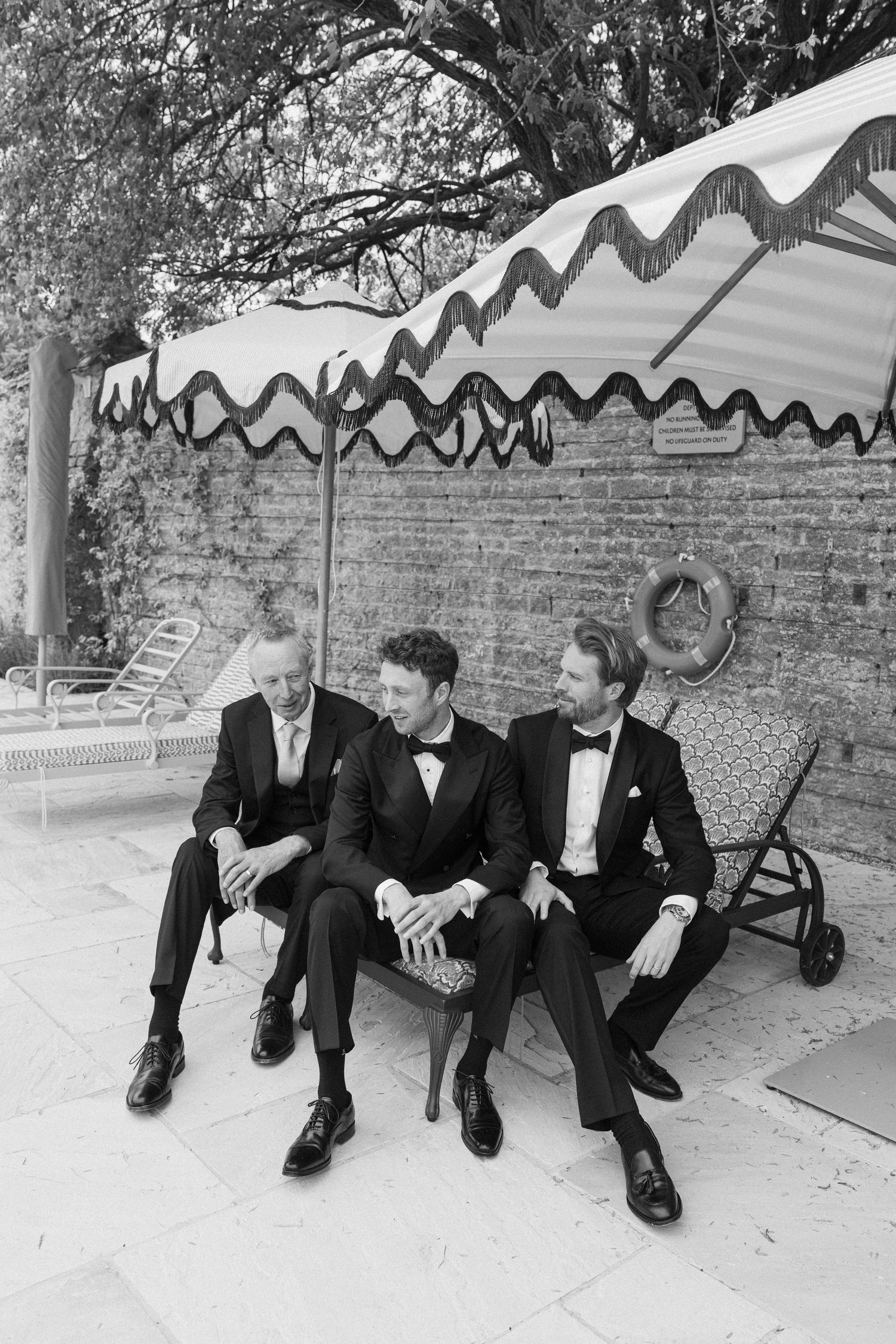Black and white photo of three groomsmen in tuxedos seated under a fringed umbrella at a wedding venue.