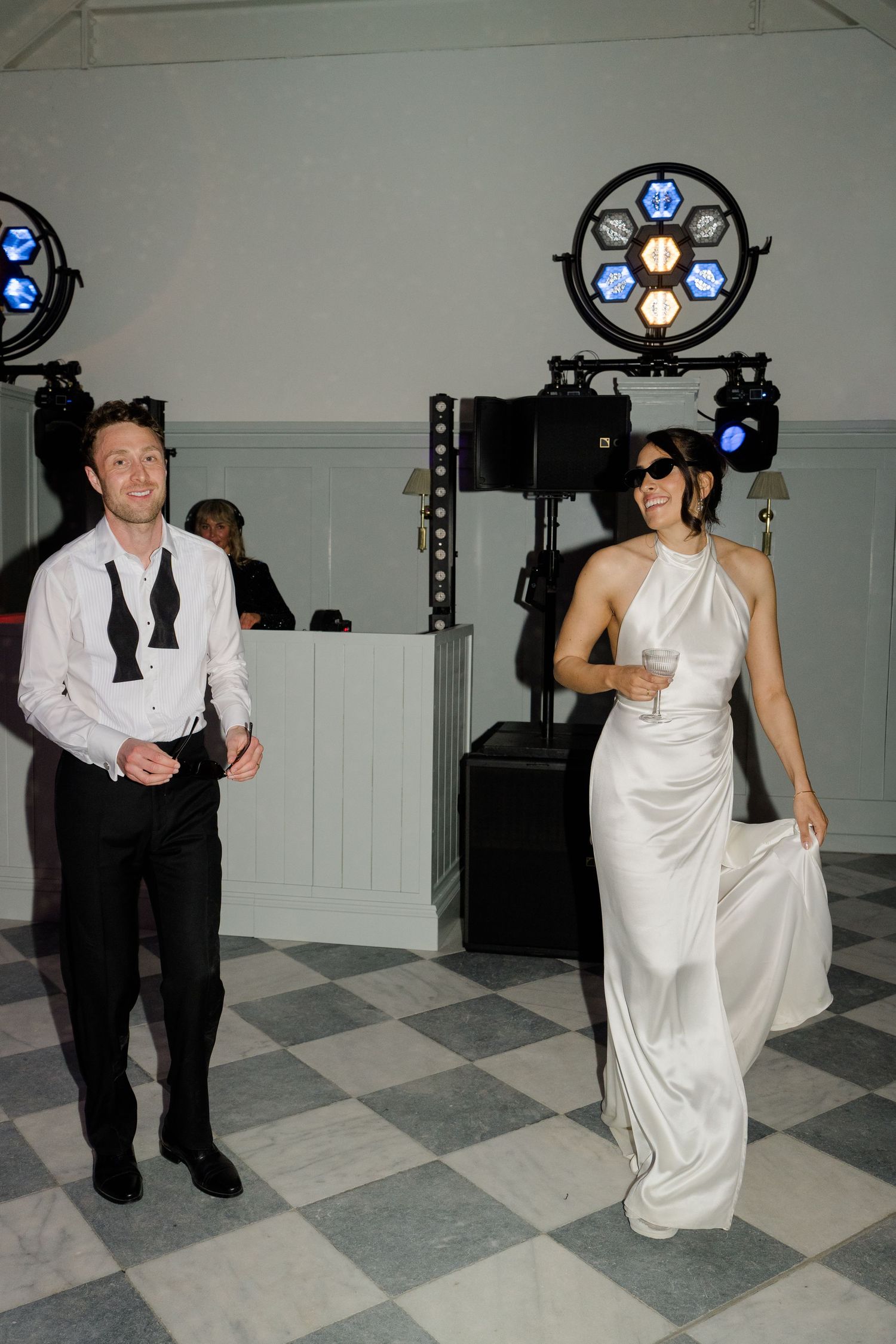 Bride in white halterneck satin gown wearing sunglasses dances with groom under colorful disco lights.